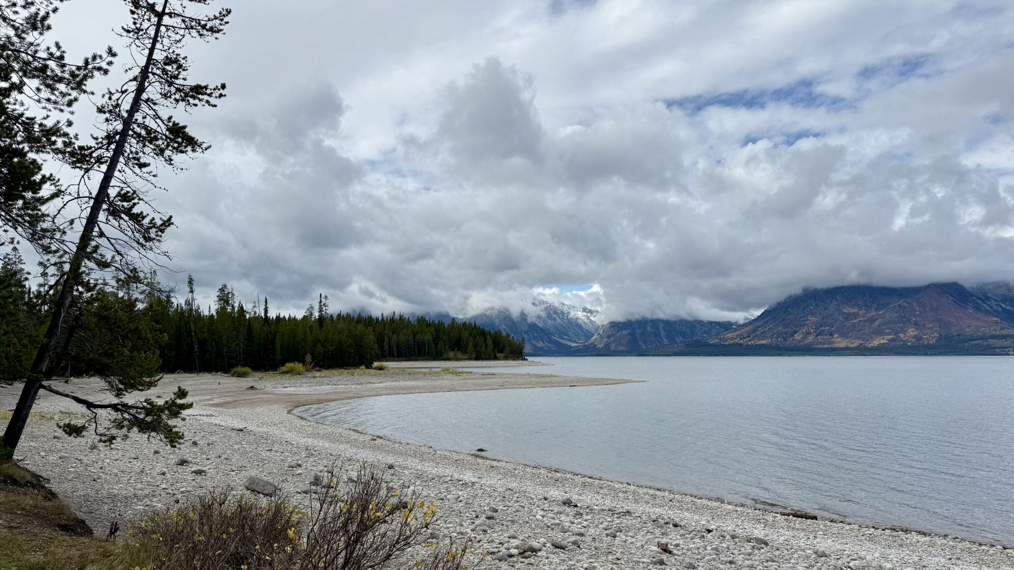Rocky beach by a calm lake, surrounded by forests and cloudy mountains. - Lakeshore Trail