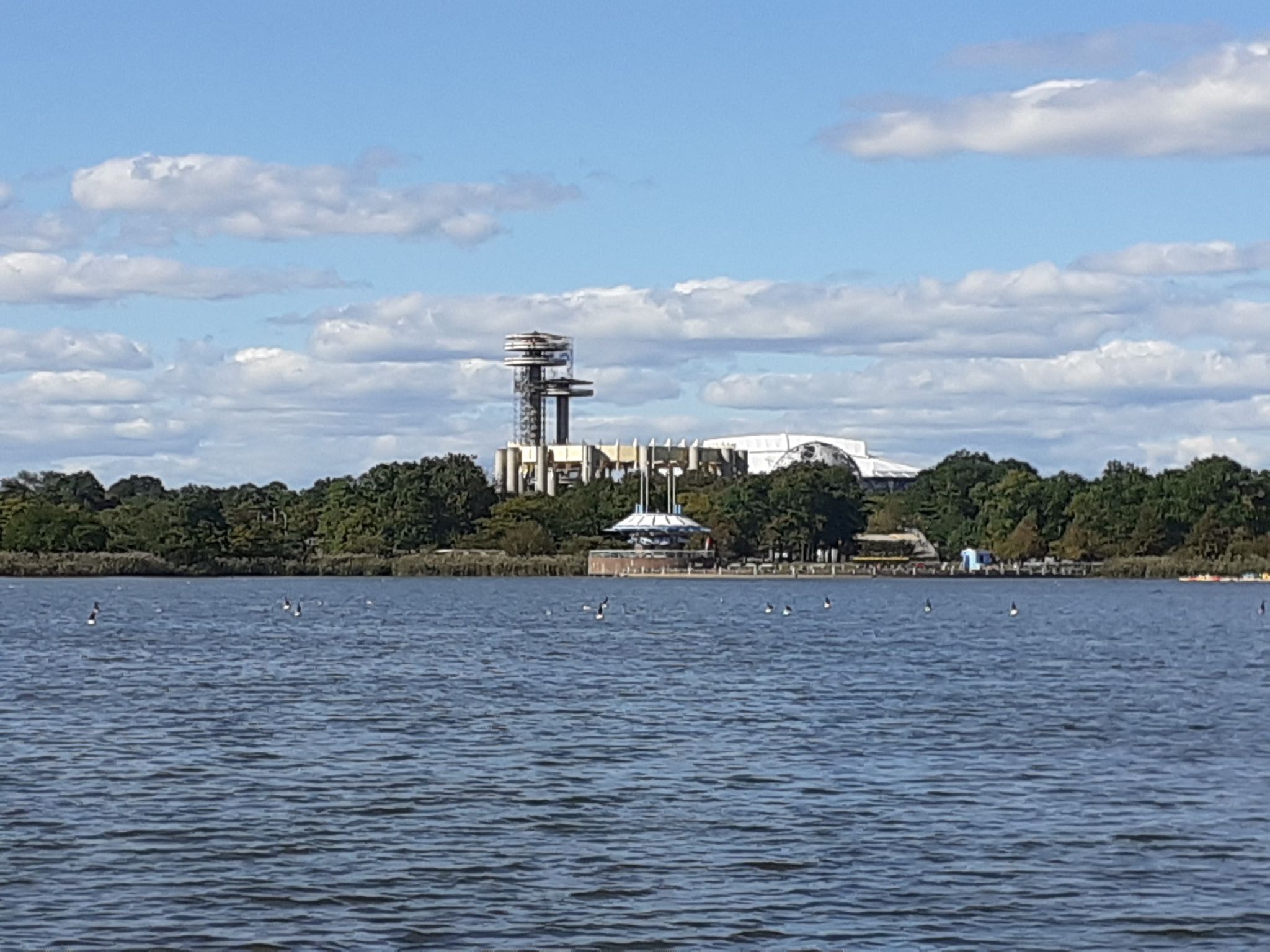 Distant buildings and a tall tower across a calm lake under a blue sky. - Flushing Meadow Park and Meadow Lake