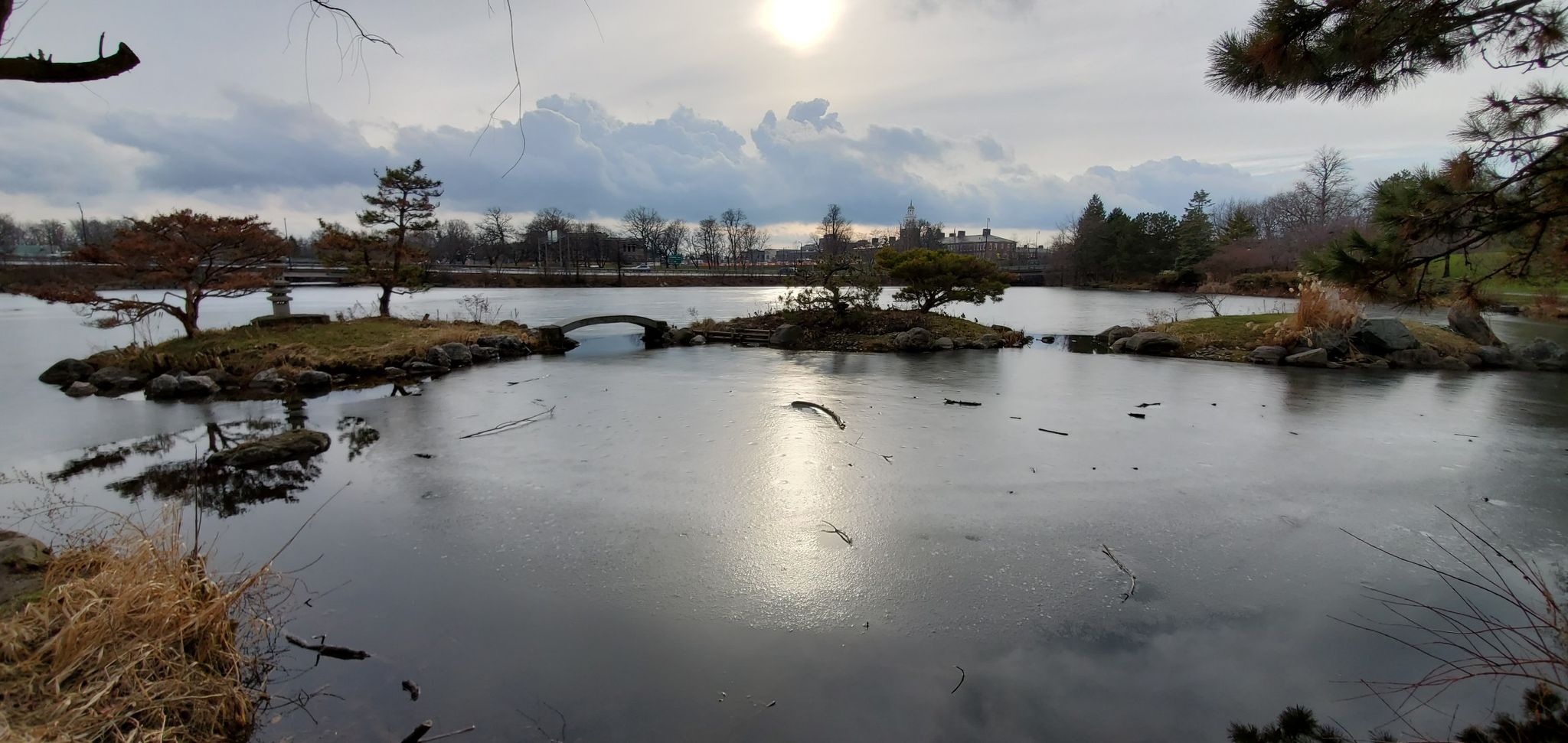 Sun shines over a partially frozen lake with small islands and bare trees. - Delaware Park to Garrison Cemetery by the International Airport loop