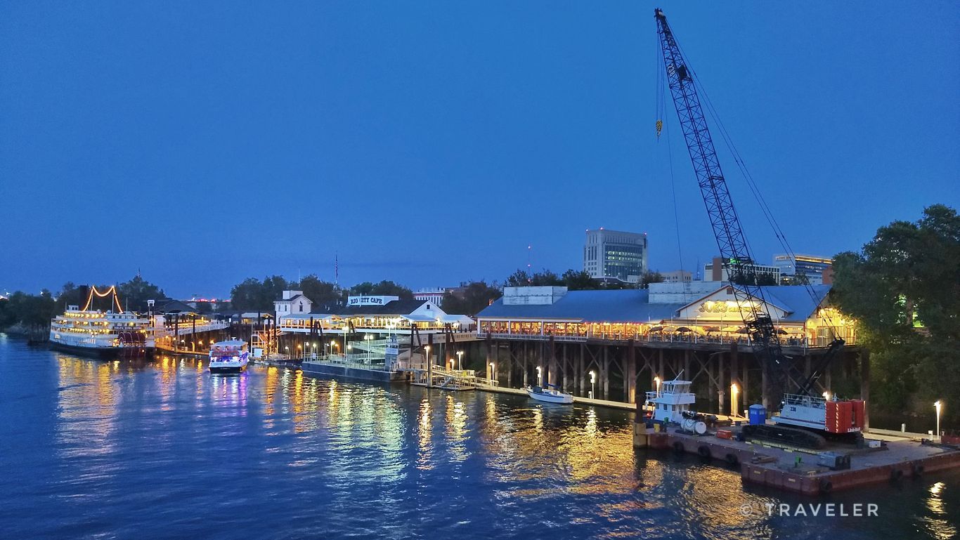 Waterfront at dusk with illuminated buildings, boats, and a large crane. - Sacramento River loop from Downtown Sacramento via Clarksburg Trail