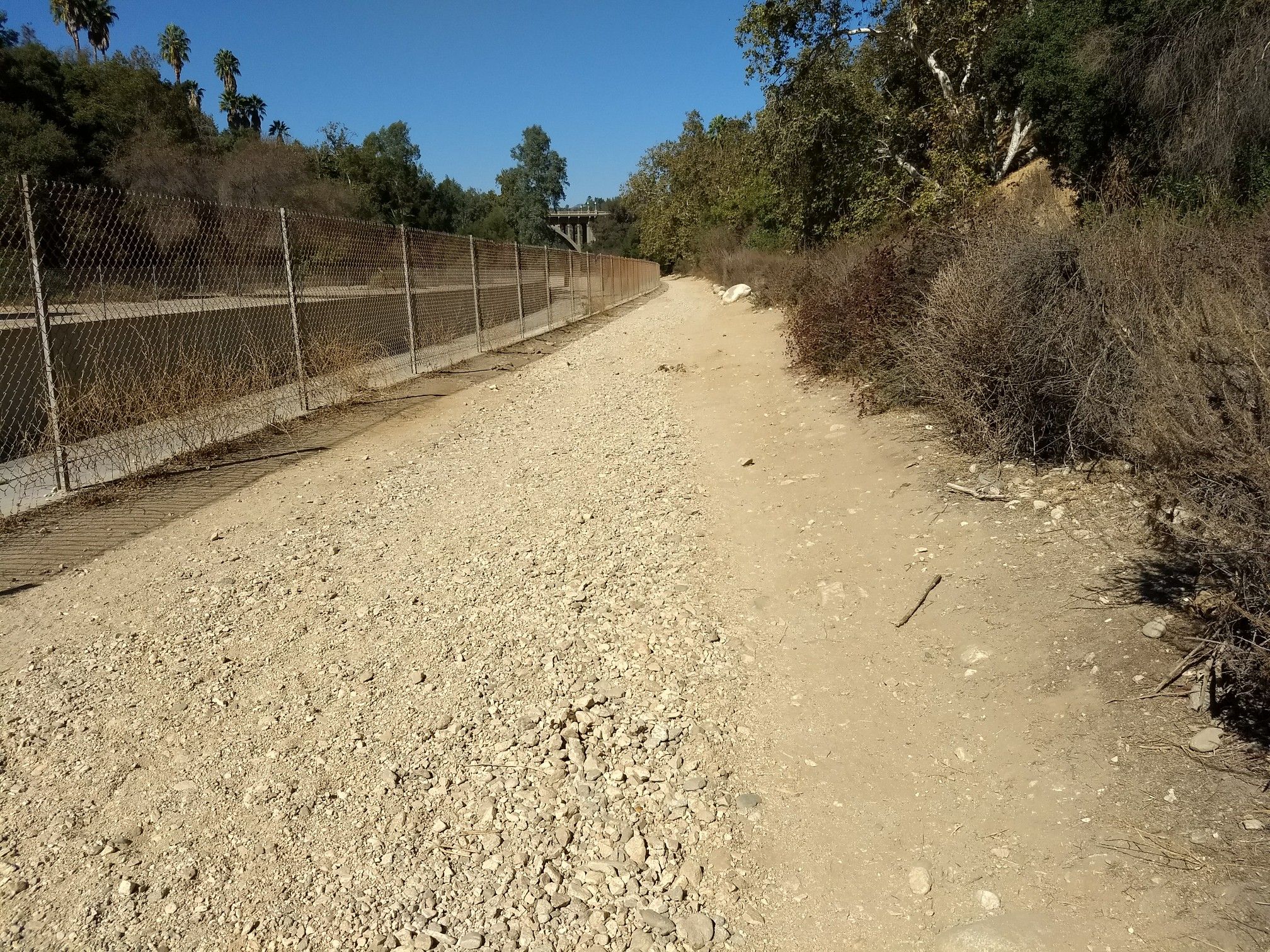 A wide gravel path runs alongside a fence and dry bushes, sunny. - Lower Arroyo Park – Arroyo Seco loop from South Pasadena