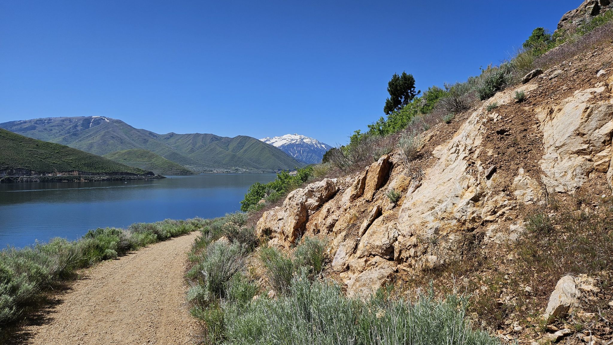 A dirt path beside a rocky hillside overlooks a lake and snow-capped mountains. - Deer Creek Trail