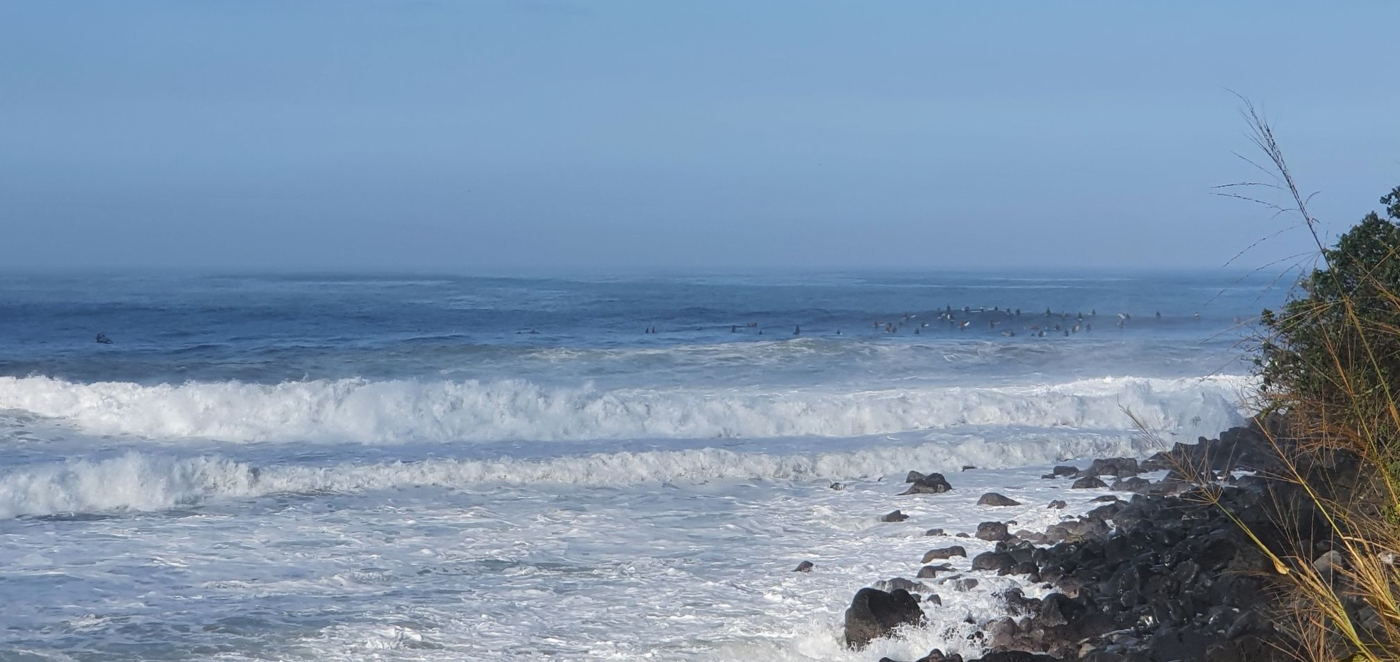 Large ocean waves crashing onto a dark rocky shore under a hazy sky. - Waimea Bay to Waimea Valley