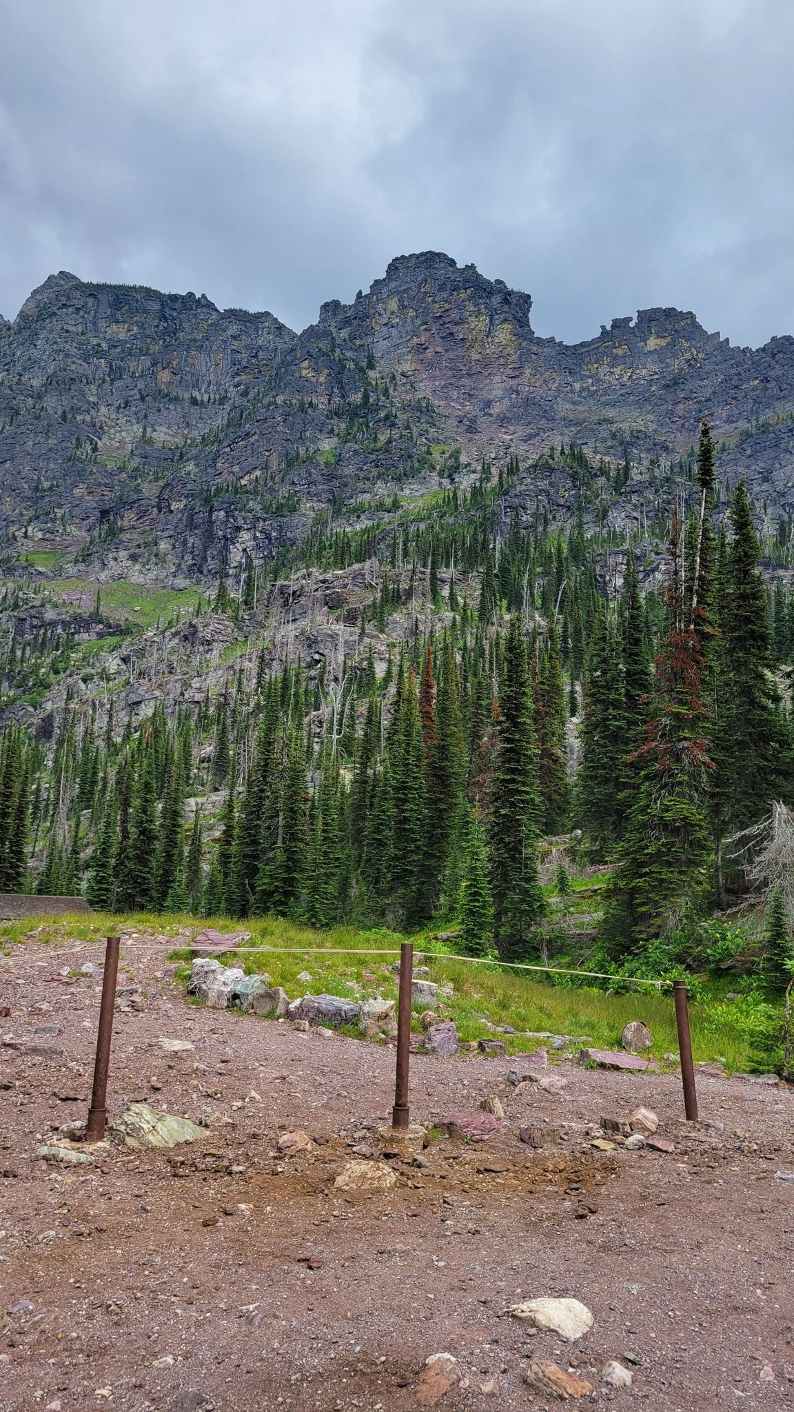 Rocky foreground with wooden posts and tape, leading to a steep, forested mountain. - Sperry Chalet