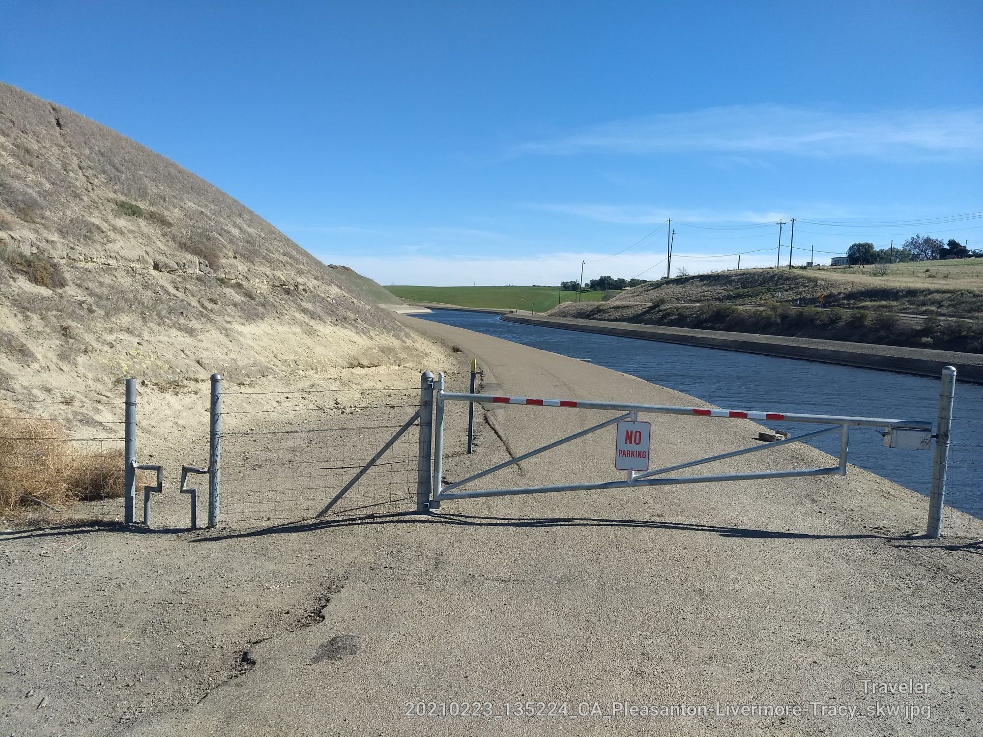 A metal gate blocks a dirt road beside a canal under a clear sky. - California Aqueduct Bikeway – Bethany Reservoir loop from Tracy