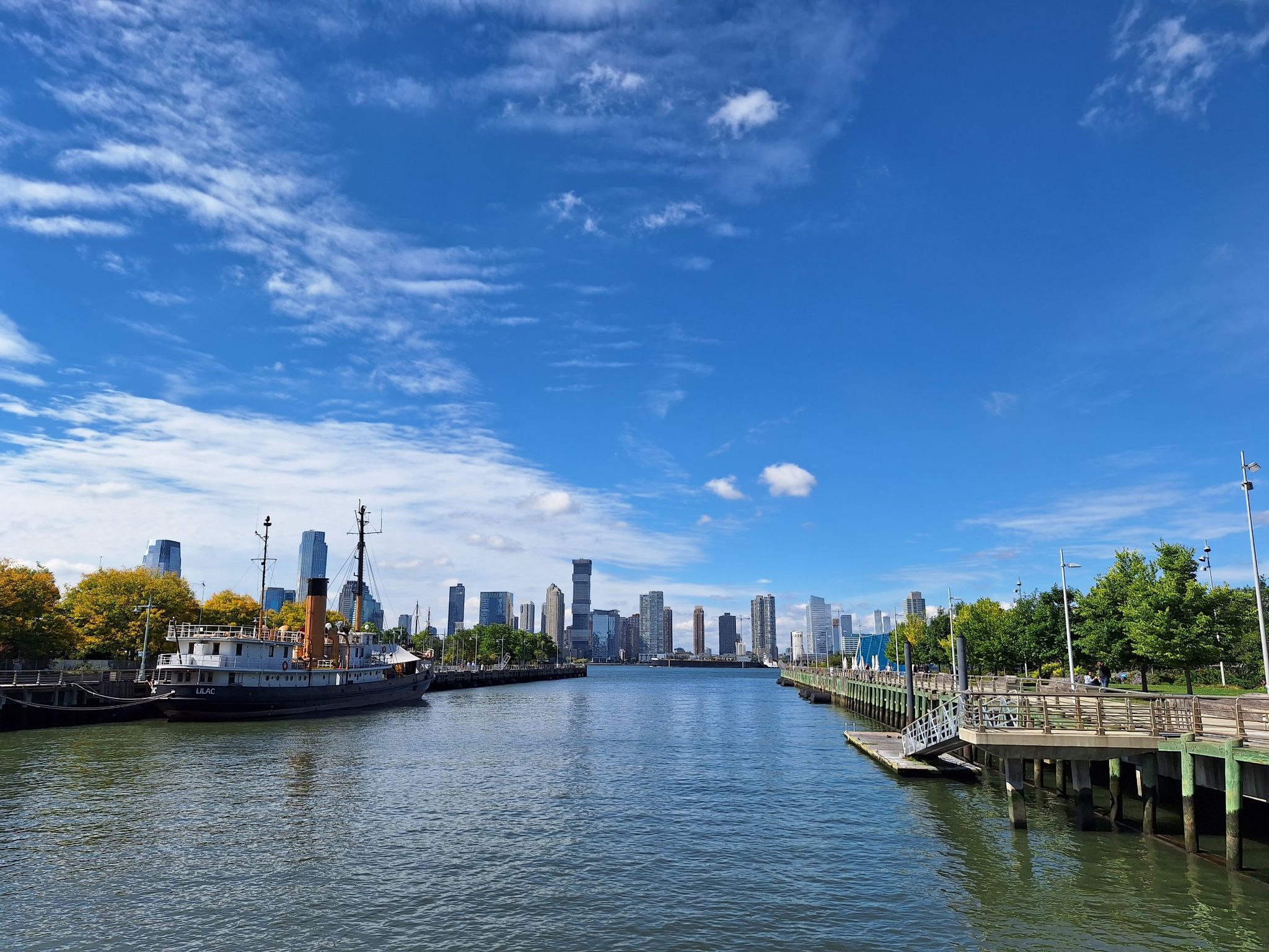A wide river with a docked boat and a distant city skyline under blue sky. - The High Line – One World Trade Center loop from Christopher Street