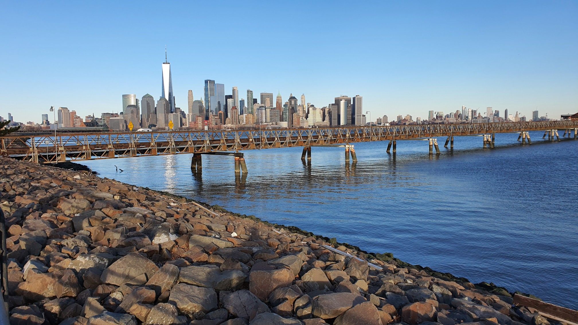 City skyline across water with a long pier, under a clear blue sky. - Hudson River Waterfront Walkway
