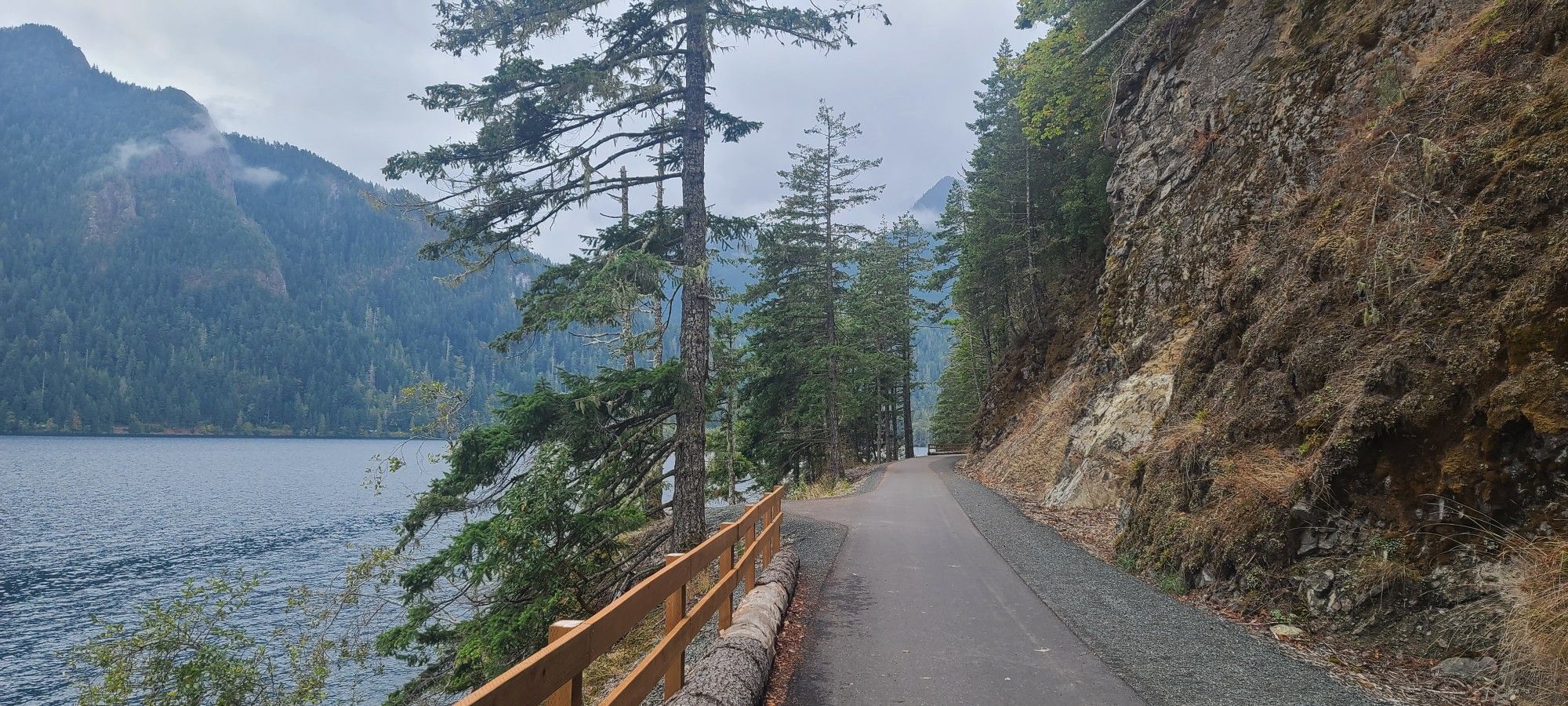 Paved path alongside a lake with mountains and evergreen trees under cloudy skies. - Spruce Railroad Trail