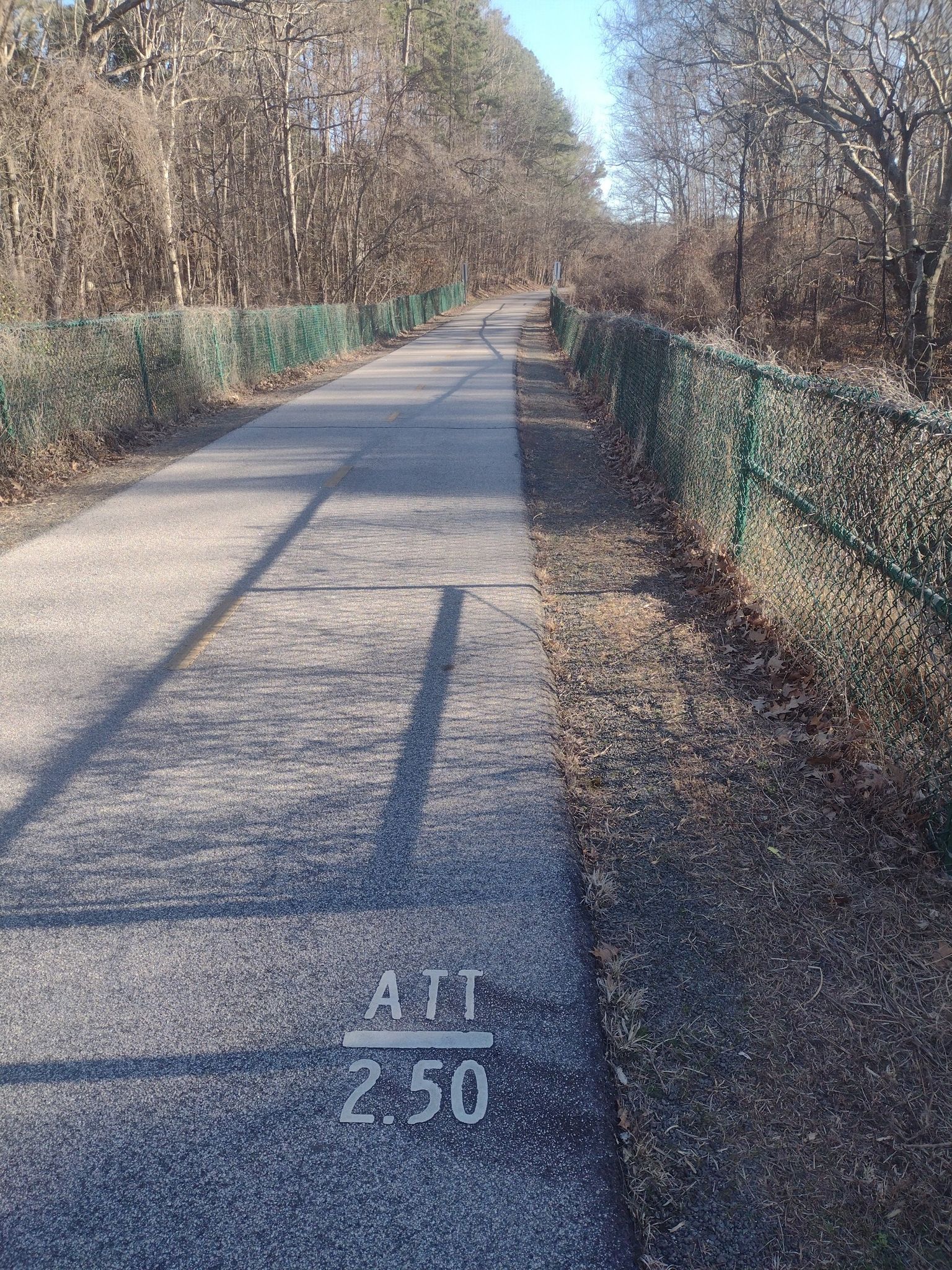 Paved path with fences, bare trees, and ground markings under sunny skies. - American Tobacco Trail