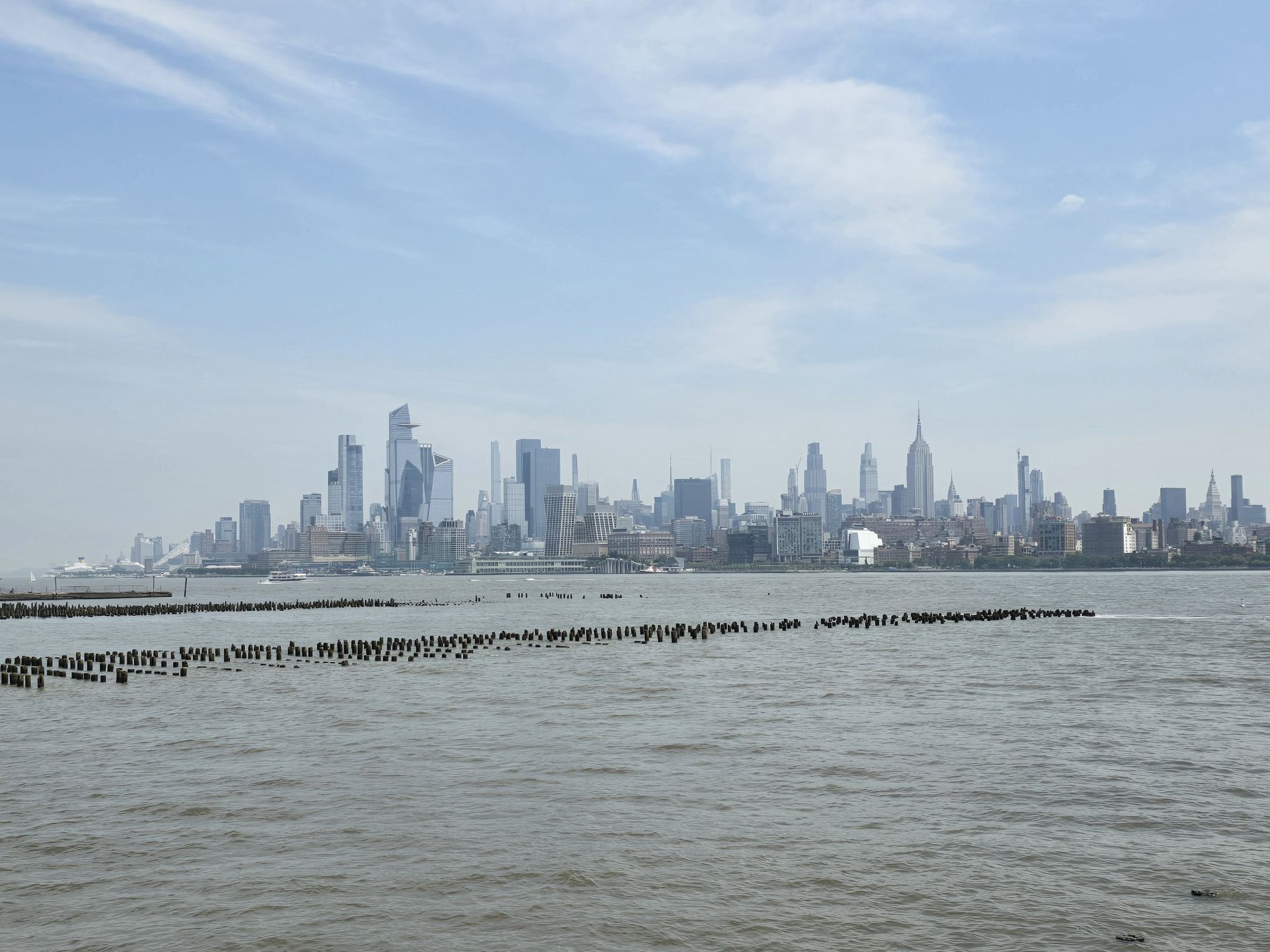 Distant city skyline across water with a long line of wooden posts. - Grove Street – LeFrak Point Lighthouse loop from Grove Street