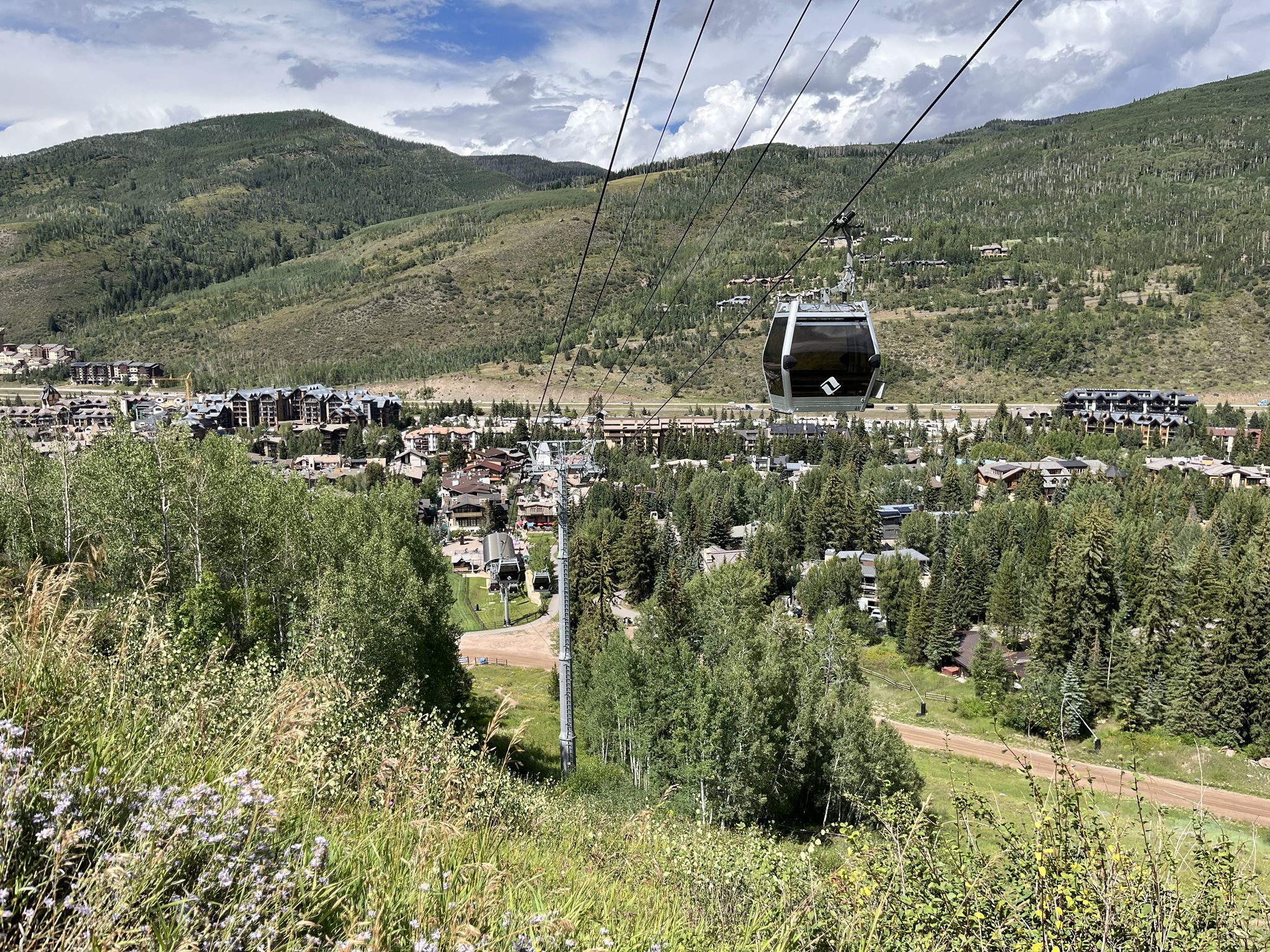 Gondola cabin over a green mountain valley with a town under a cloudy sky. - Gore Creek Path – Vail loop from Vail