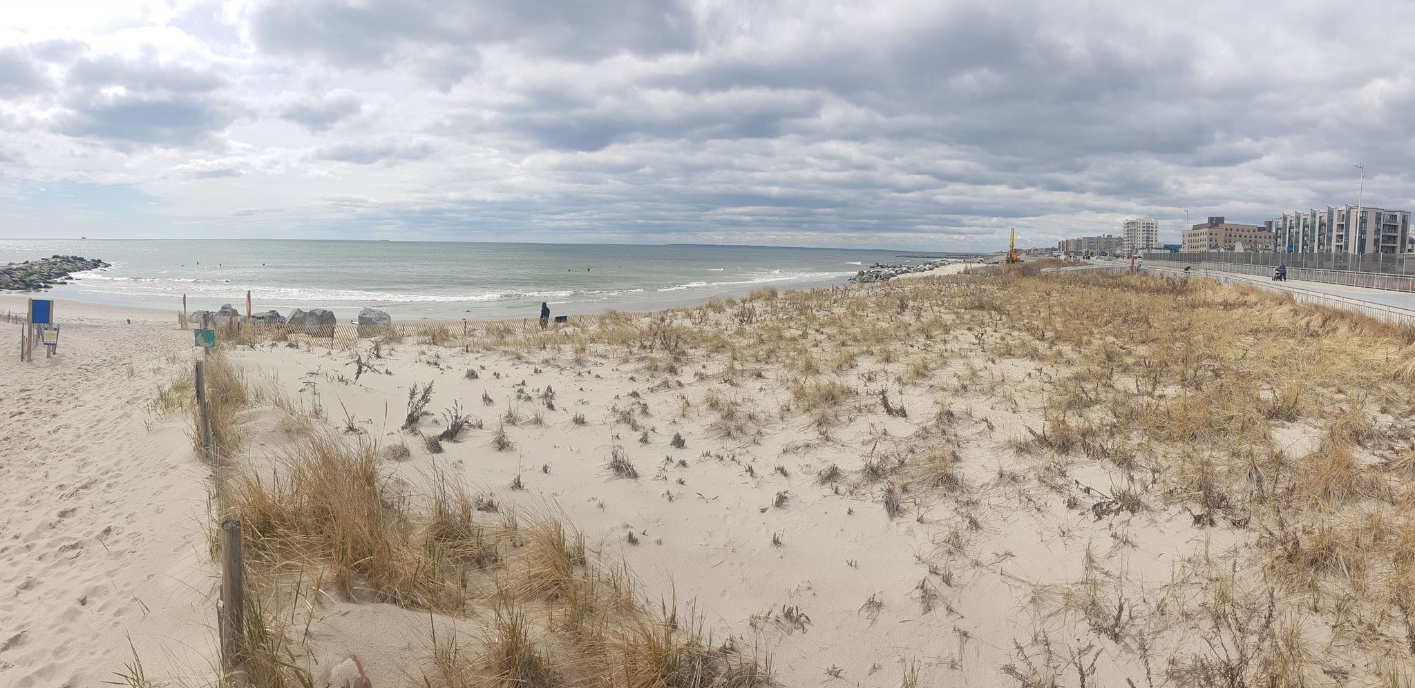Overcast panoramic beach with sand dunes, ocean, and distant buildings. - Rockaway Beach Boardwalk – Rockaway Beach Boardwalk loop from Beach 67th Street
