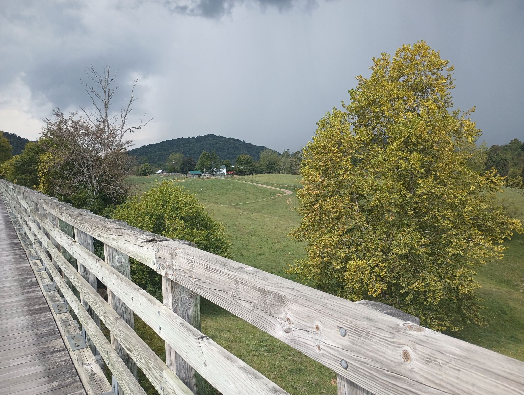 A wooden bridge railing overlooks a green valley with distant hills under a cloudy sky. - Virginia Creeper Trail