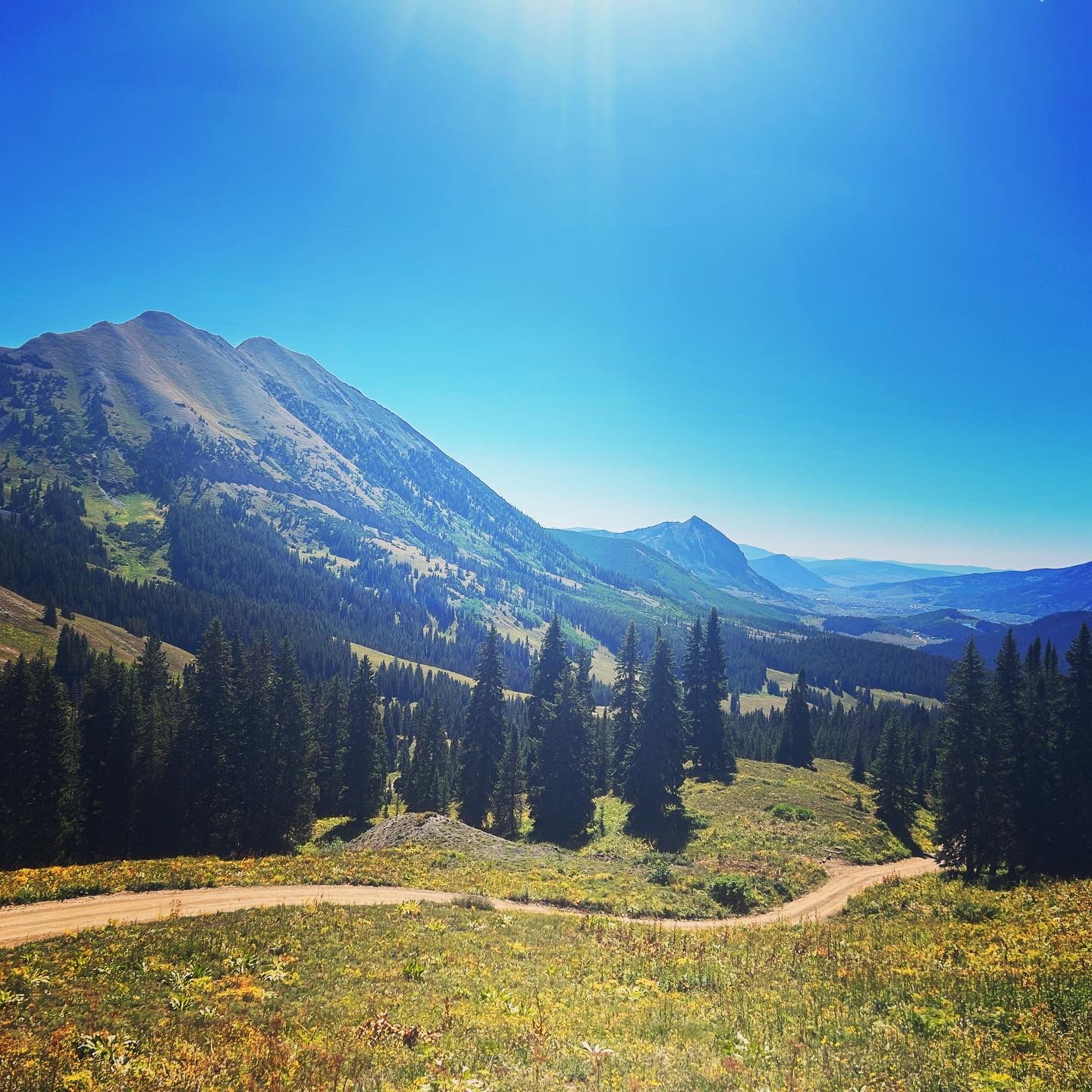 Sunny mountain landscape with a dirt road winding through a forested valley. - Crested Butte Downtown loop from Upper Loop