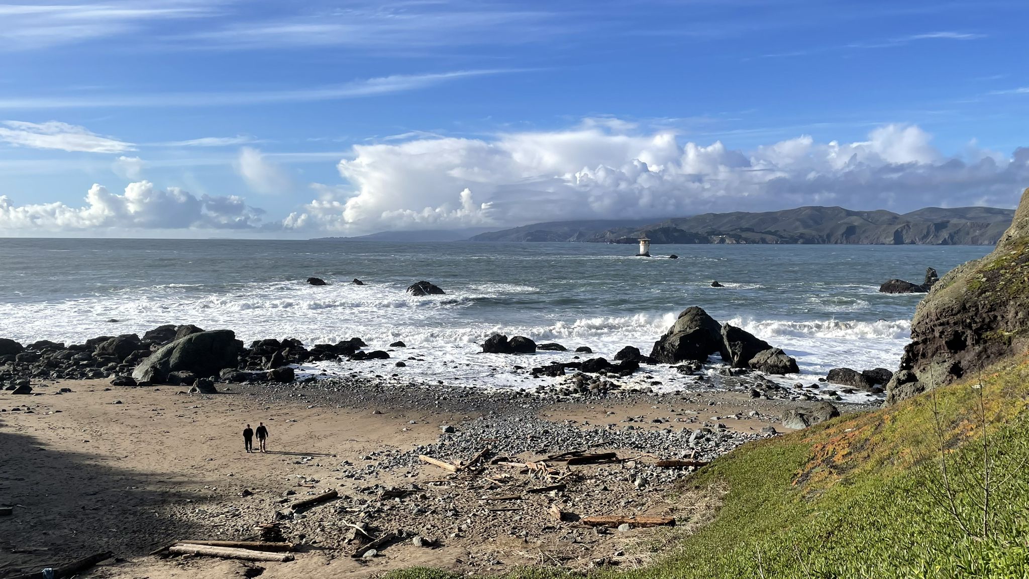 Two people walk on a rocky beach with crashing waves and distant hills. - Lands End Beach – Coastal Trail - Land's End loop from Judah Street & 9th Avenue