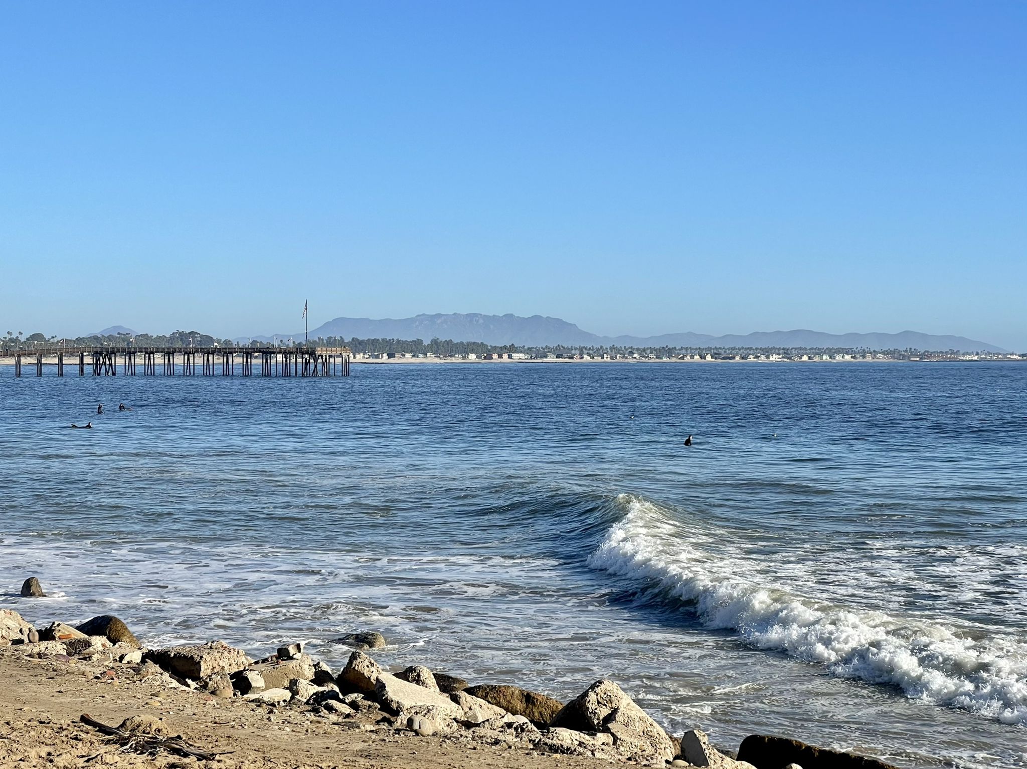 Ocean with small waves, distant surfers, and a long pier under a clear blue sky. - San Antonio Creek Bridge – Ventura Pier loop from Ventura