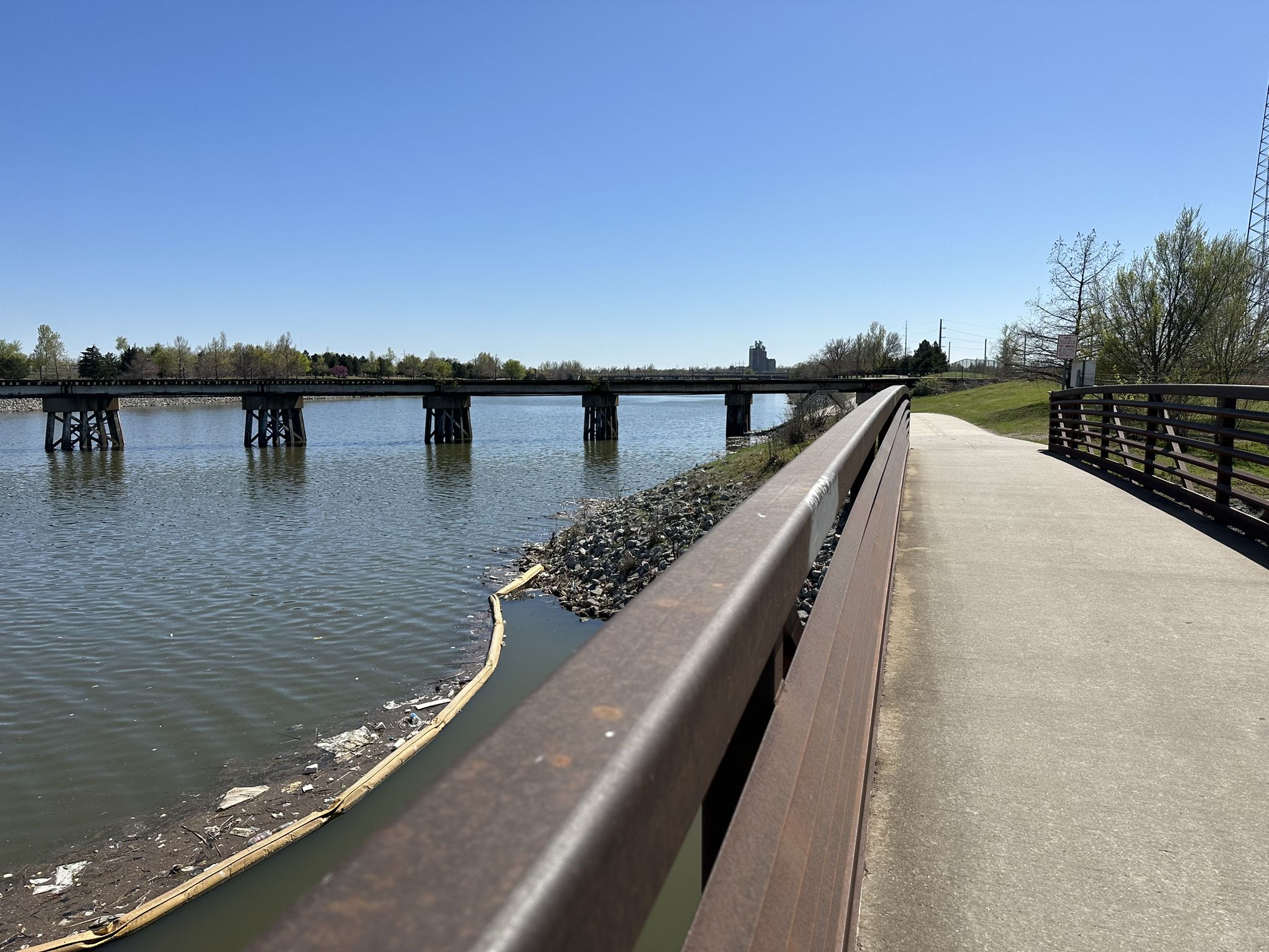 Paved path and railing alongside a lake with a long bridge in sunlight. - Oklahoma Land Run Monument – Regatta Park loop from Oklahoma City