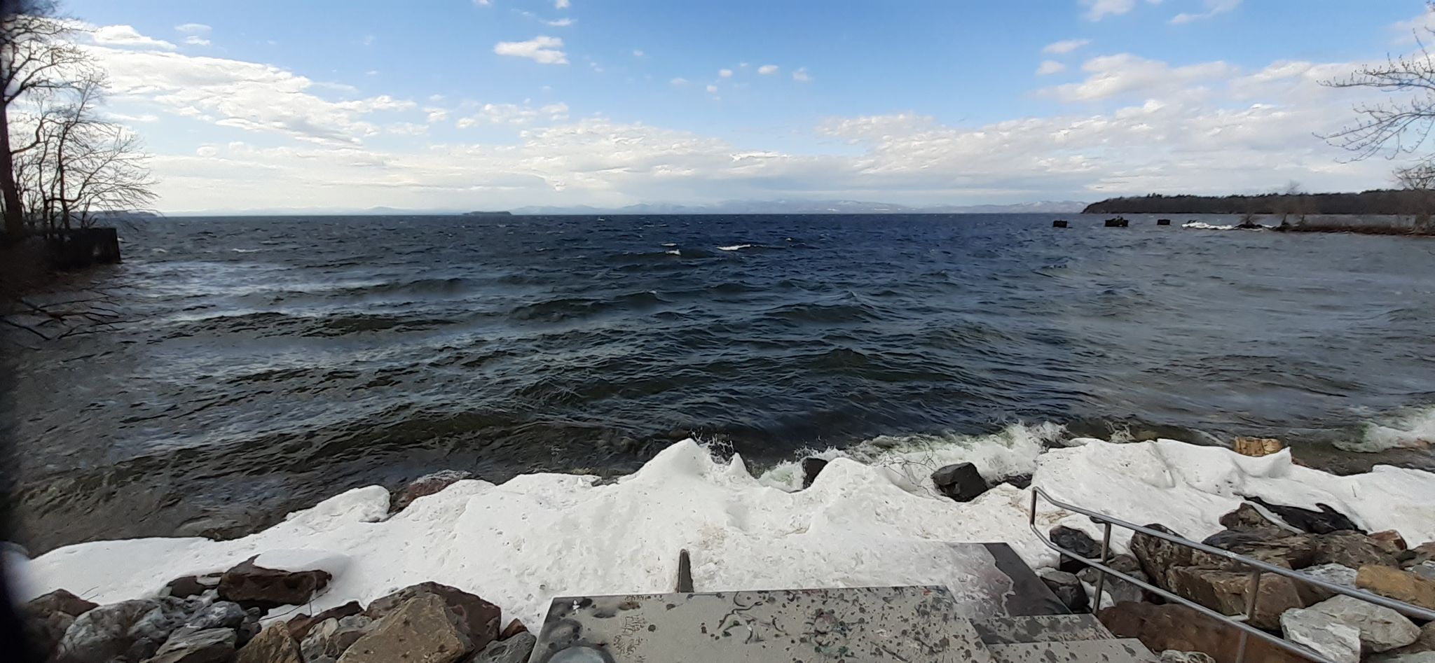 Wavy lake with snow and rocks on the shore, distant mountains, under a cloudy sky. - Granite Steps – Colchester/Burlington bridge loop from Burlington