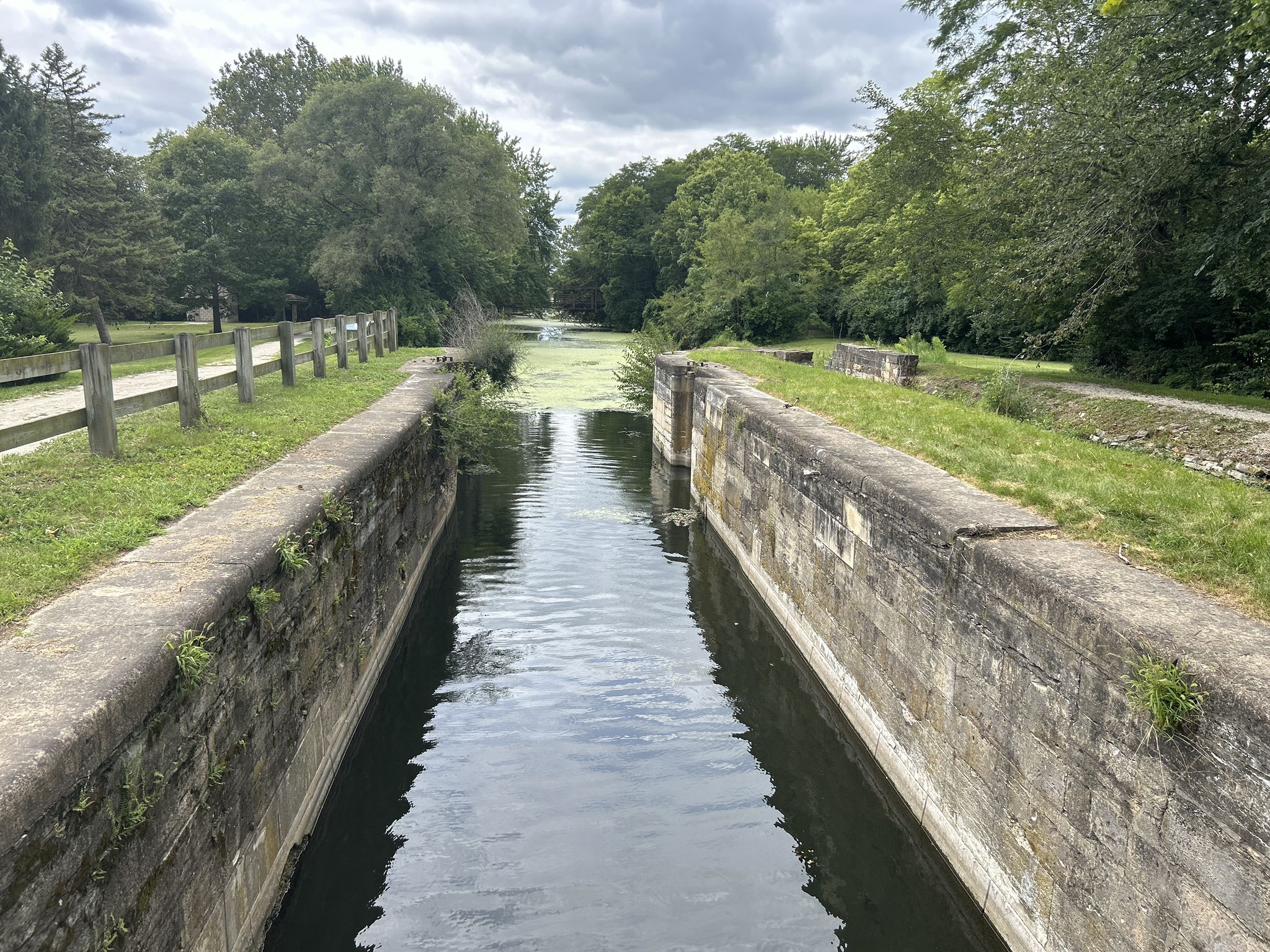 Narrow canal with concrete walls, green algae, and distant trees under cloudy sky. - Bike loop from Morris