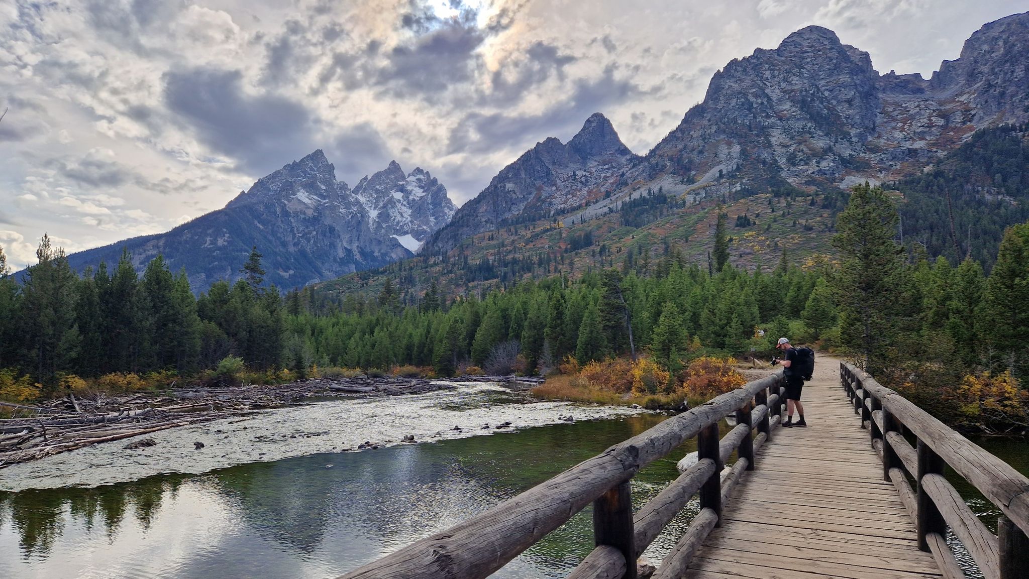 Person on a wooden bridge overlooks a wide river, forest, and rugged mountains. - String Lake Footbridge – String Lake loop from Jackson Lake Lodge