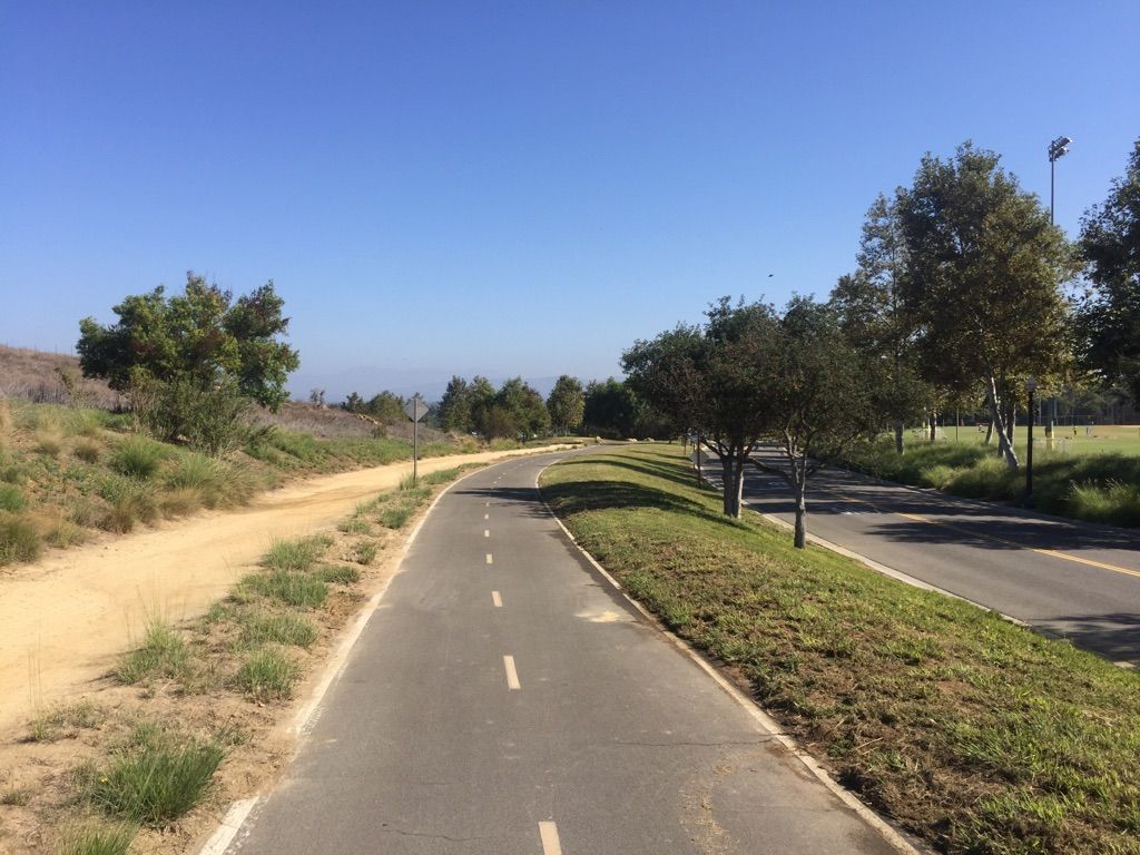 Paved bike path and dirt trail alongside trees under a clear blue sky. - Shady Canyon Trail loop from Quail Hill
