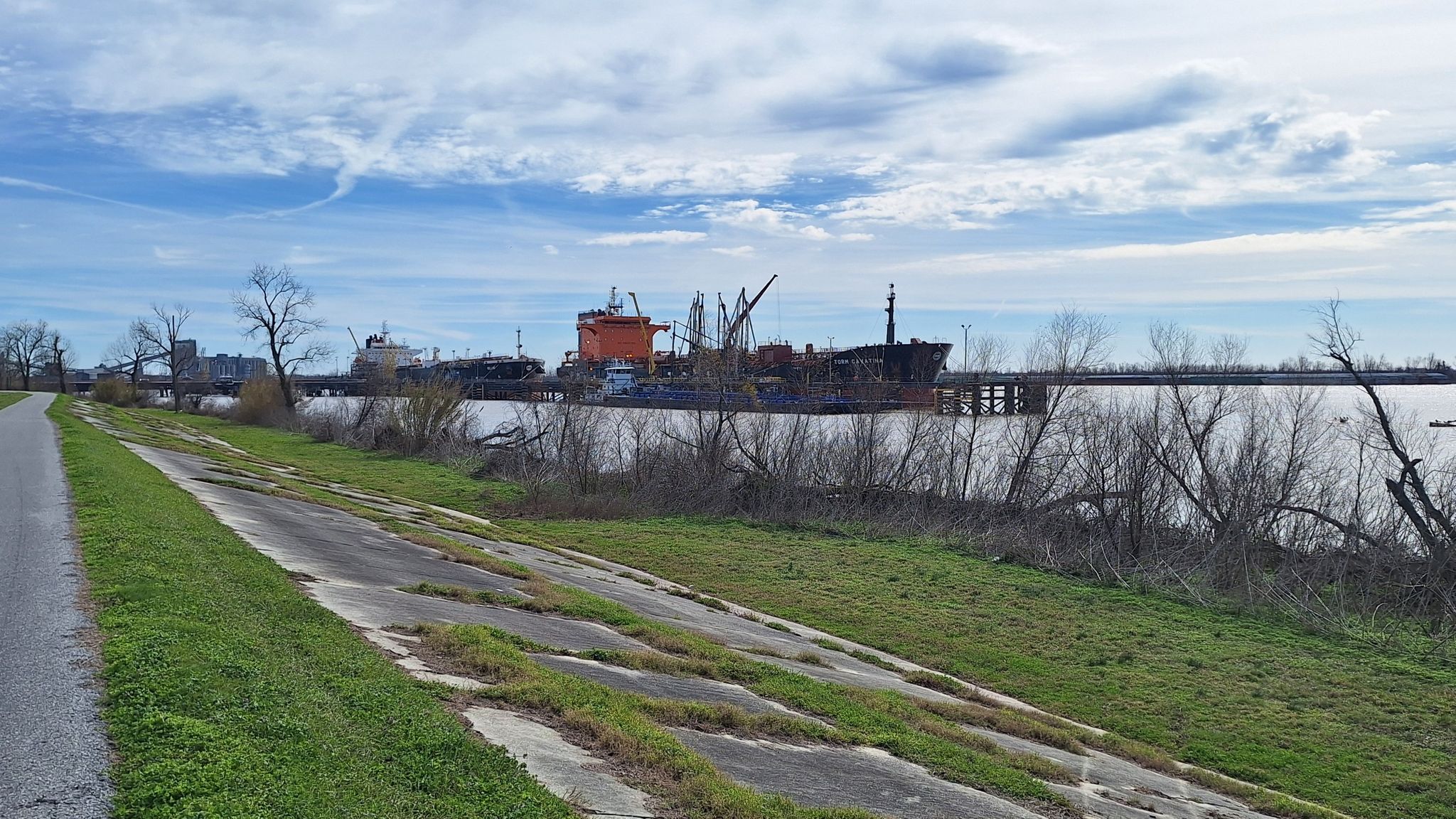 A paved path runs alongside water with large industrial ships in the distance. - Roadbike loop from Kenner