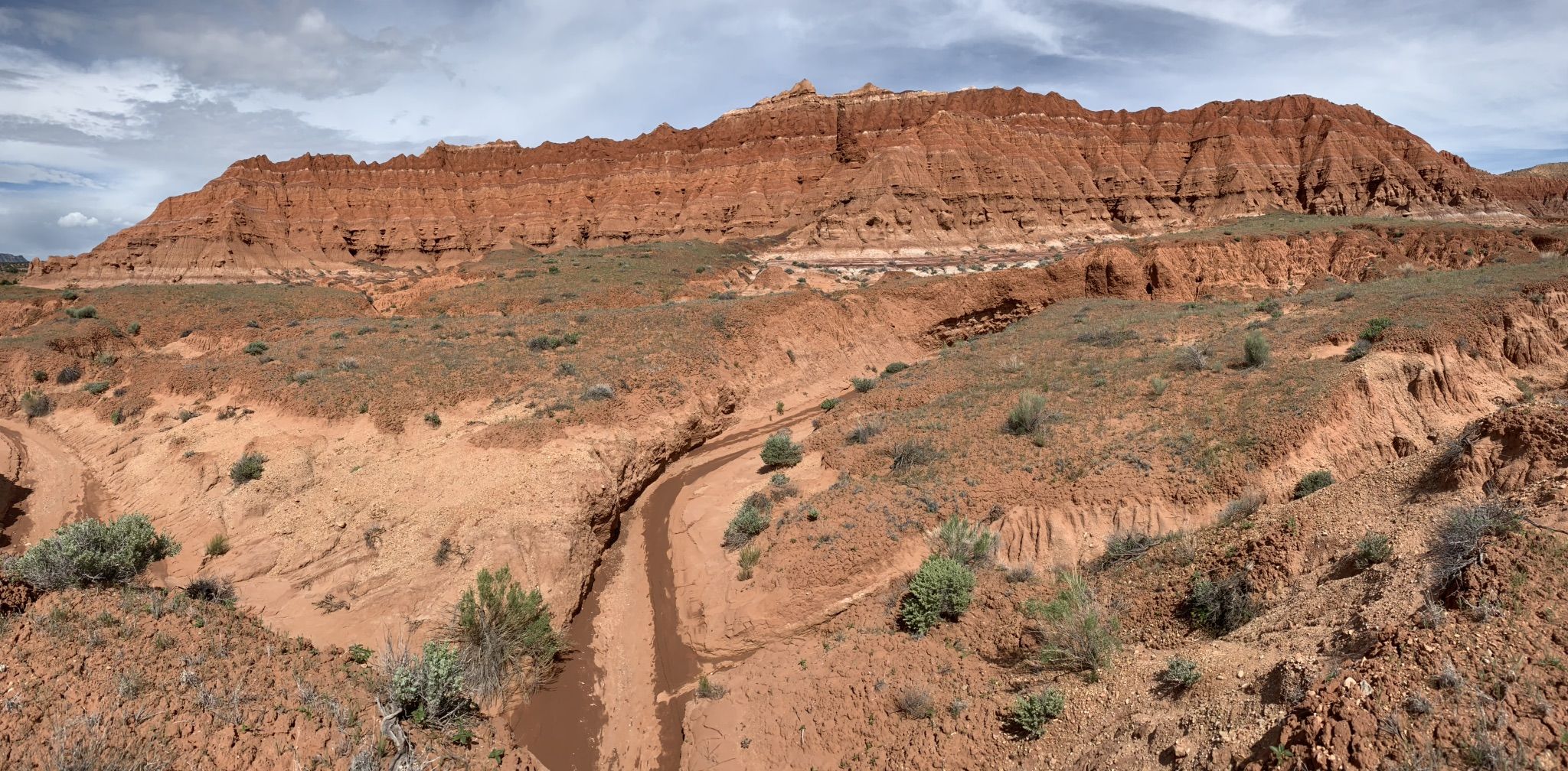 Wide view of a dry wash leading to towering red rock cliffs under a cloudy sky. - Trail To Aetschi Baetschi Tower