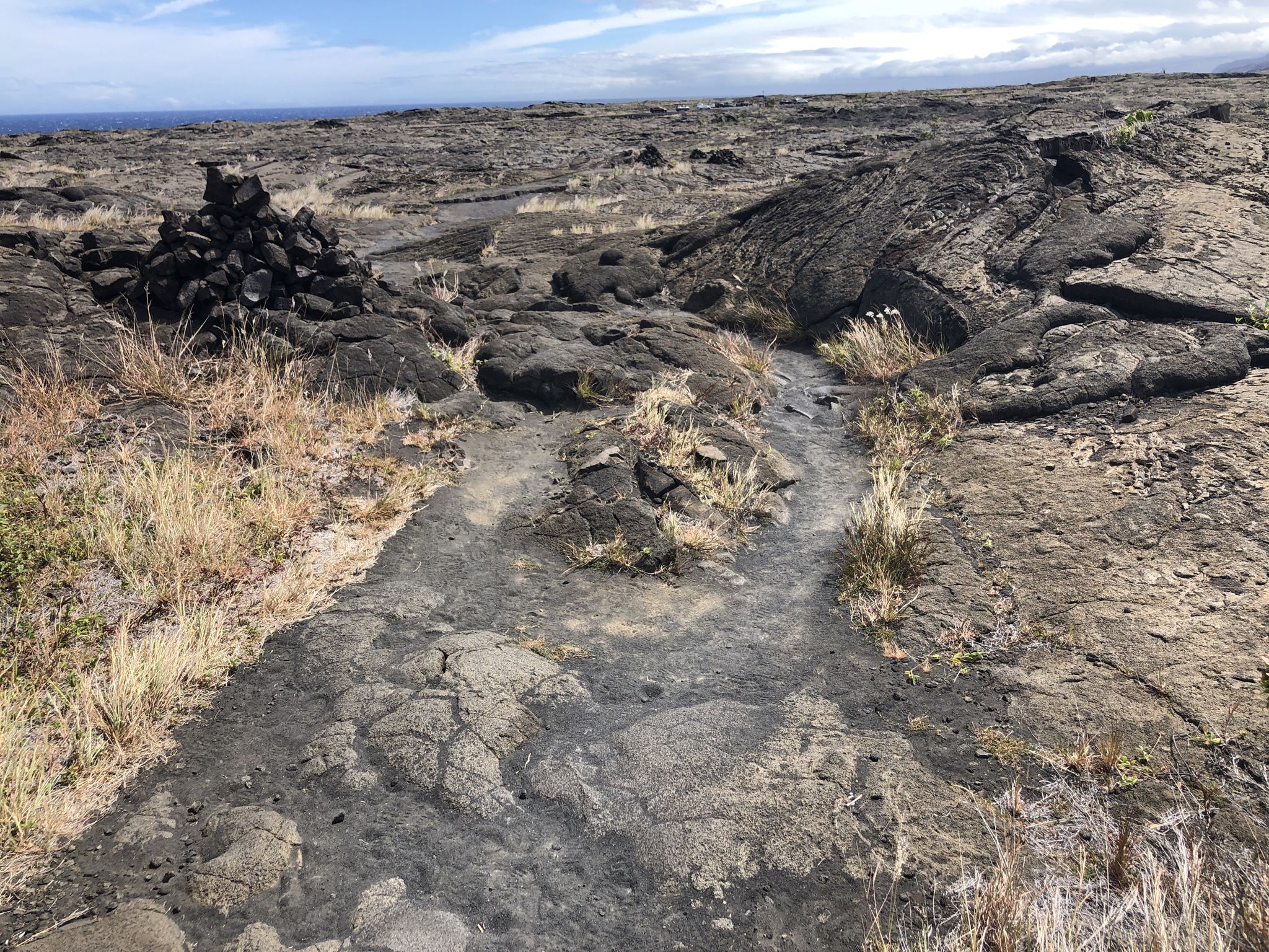 Rocky path marked by a cairn, sparse grass, and distant ocean. - Kealakomo Overlook to Pu‘uloa Petroglyphs via Chain of Craters Road — Hawaiʻi Island