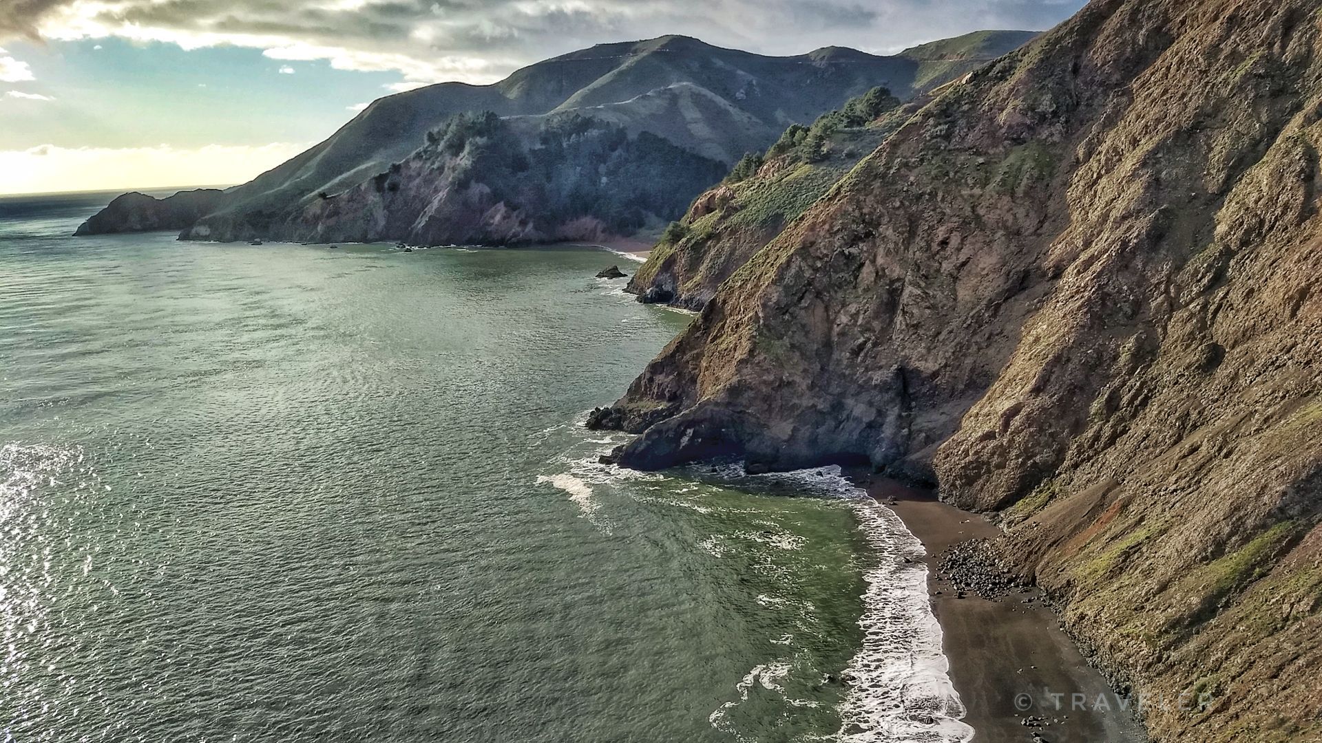 Rugged coastline with steep cliffs, a small beach, and waves. Overcast. - View of the Golden Gate Bridge – Golden Gate Bridge loop from Alcatraz Island