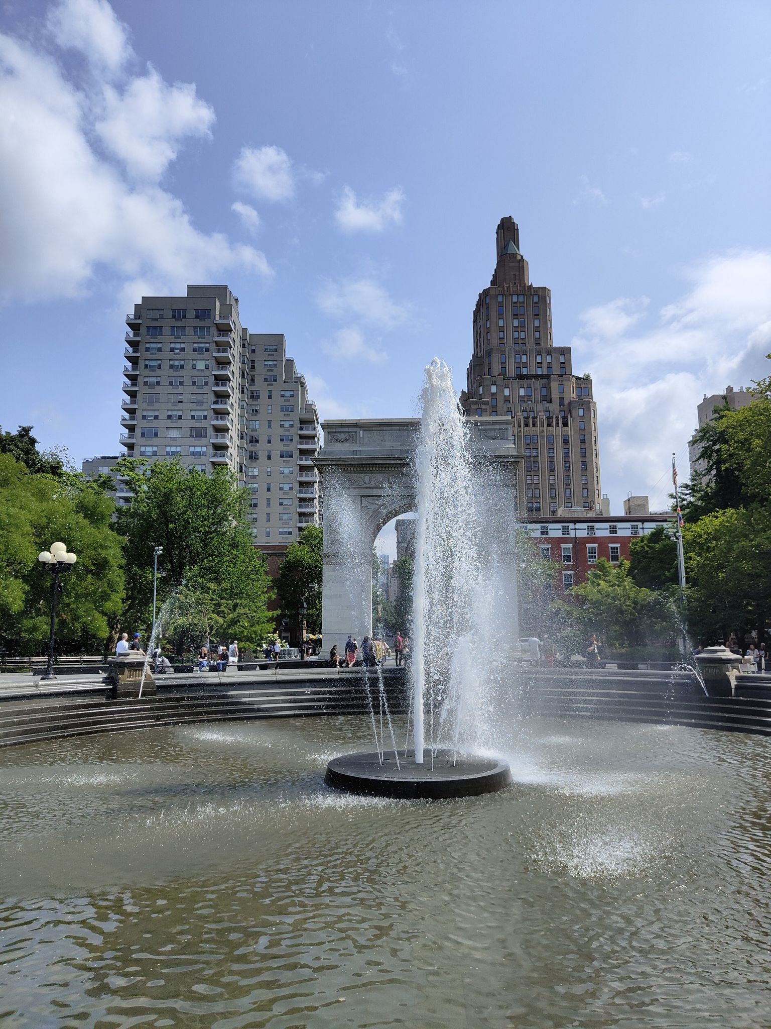 Daylight view of a fountain spraying water in front of a large archway and buildings. - Washington Square Park Loop