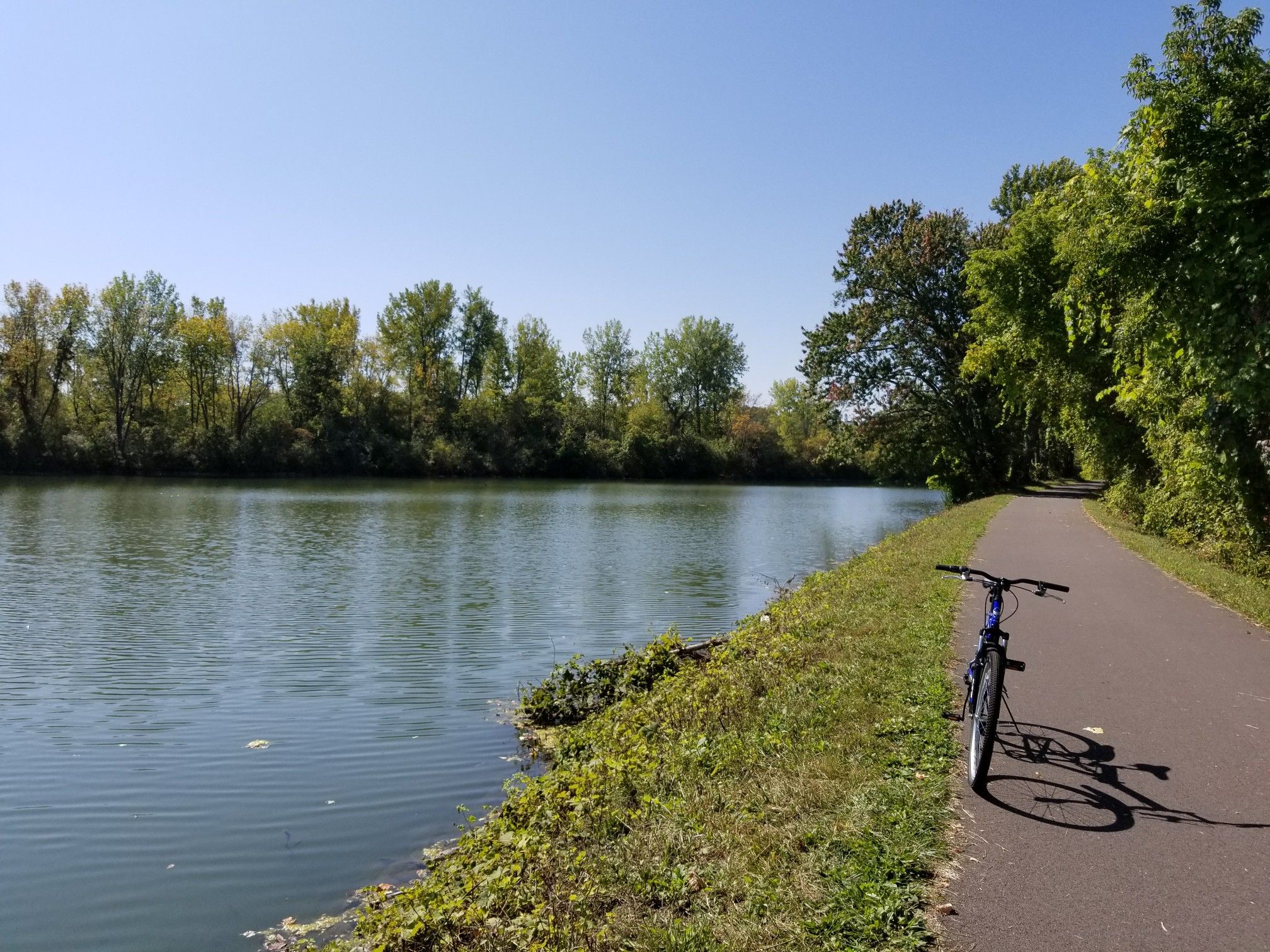 Blue bicycle parked on a paved path beside a wide river, sunny day. - Erie Canalway Trail – Erie Canalway Trail loop from Syracuse
