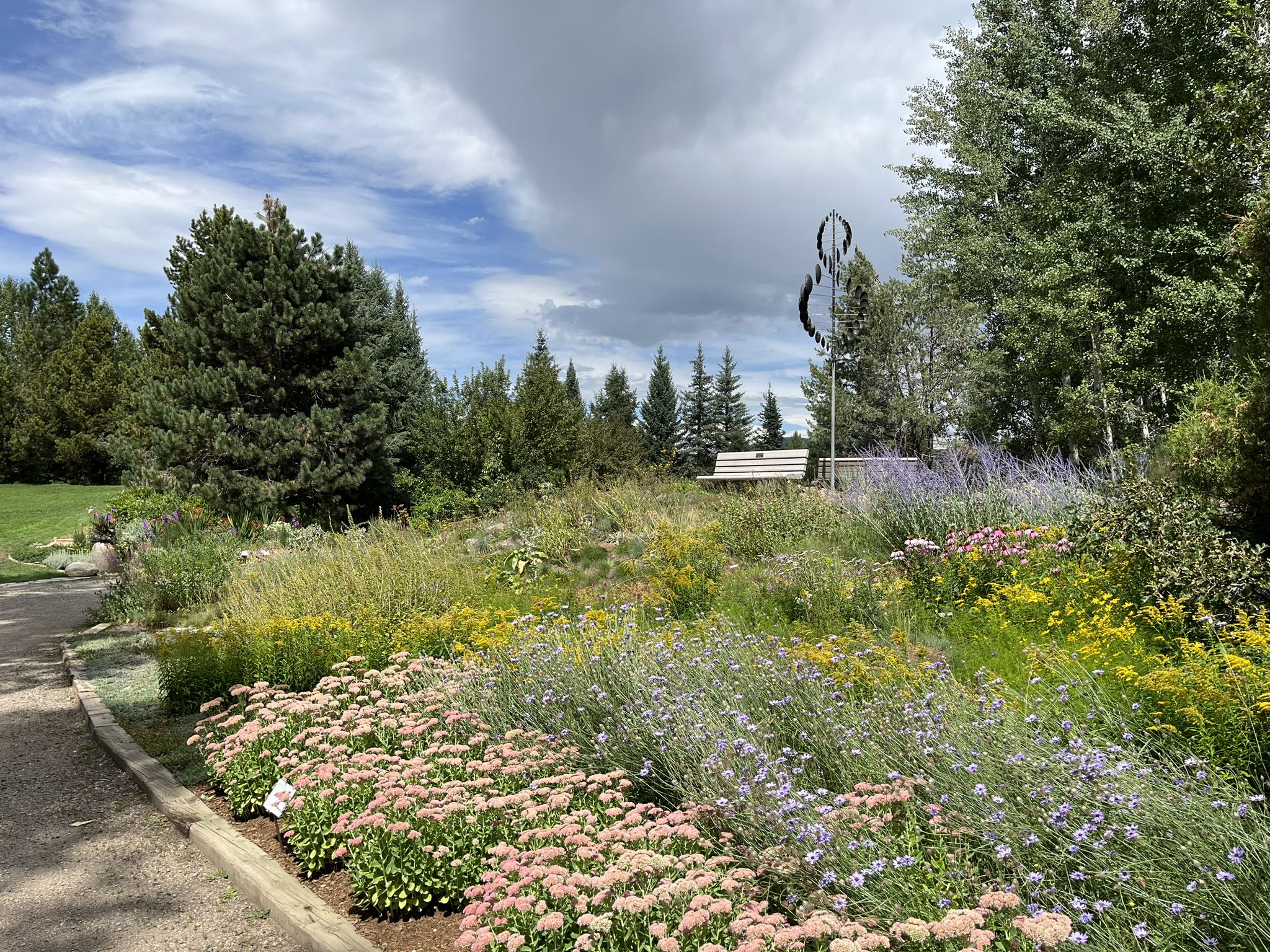 Lush garden with colorful flowers, trees, a bench, and sculpture under a cloudy sky. - Yampa River Core Trail