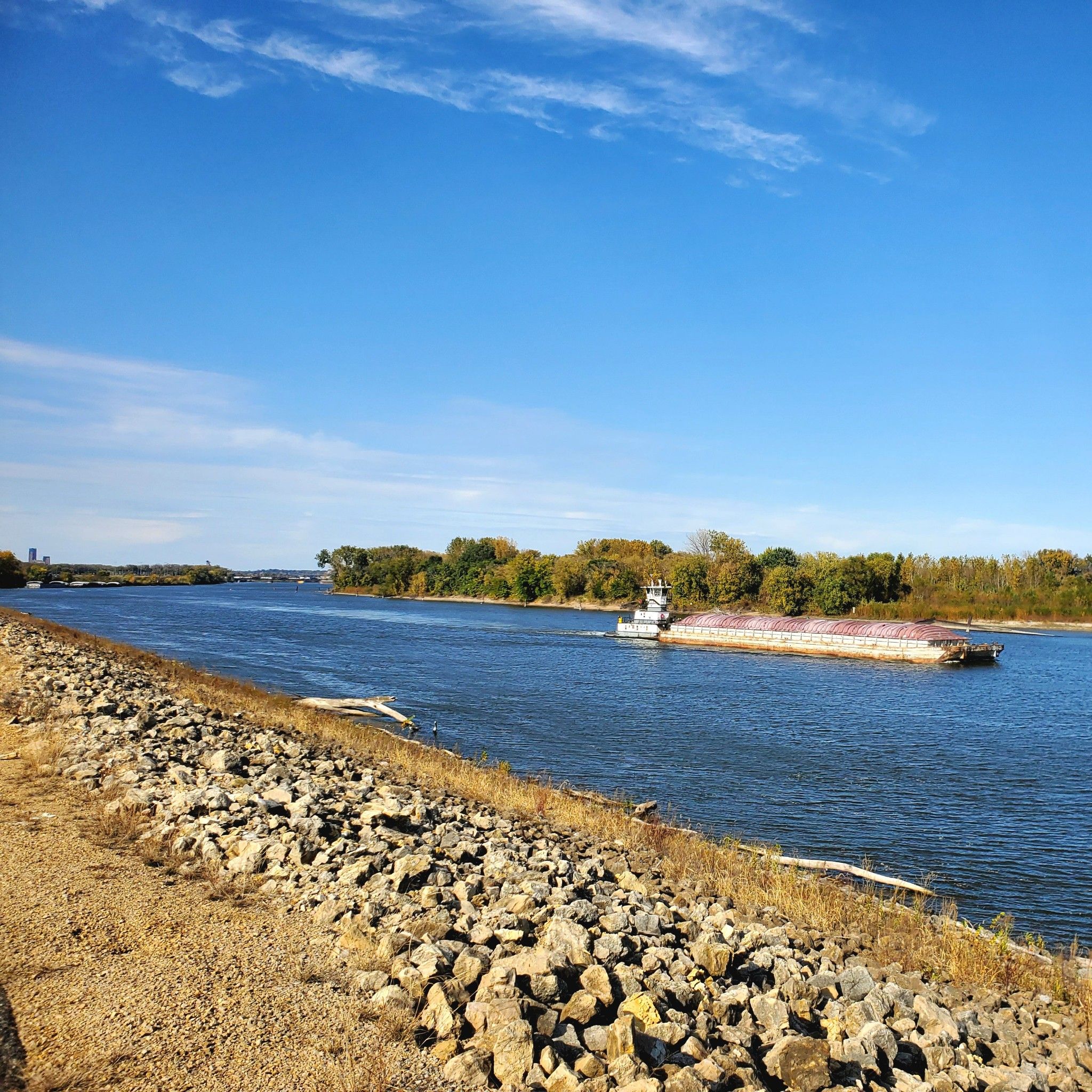 Large barge moving along a wide river with a rocky shore under blue sky. - Mississippi River Greenway