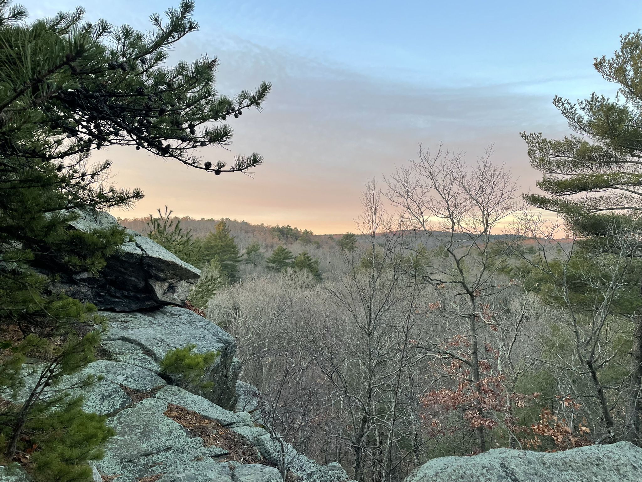 Rocky foreground frames a forest vista under a soft, colorful sky. - Saint Moritz Pond – Nahanton Hill loop from Granite Railway Incline