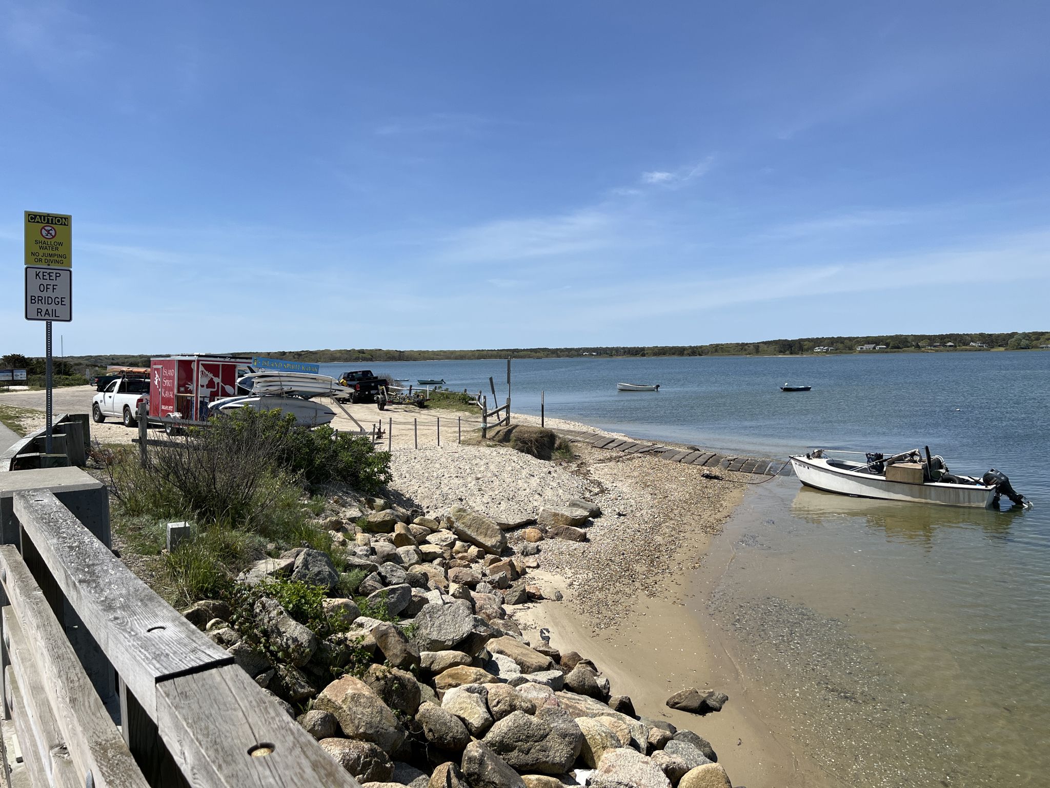 Small boats on a rocky beach next to a road and calm water. - View of the Marshland loop from Trade Wind Airport