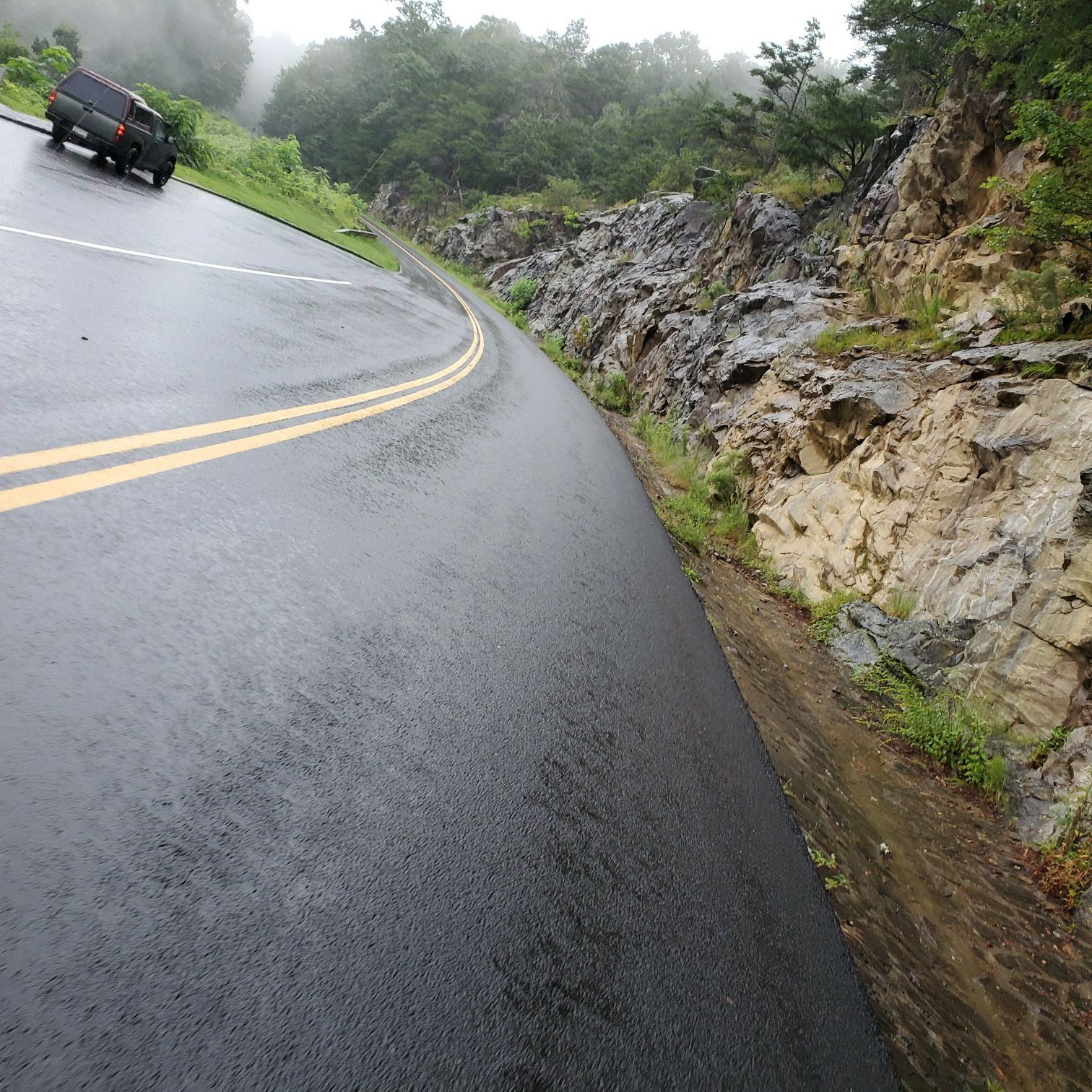 A wet, winding road with a car, rocky hillside, and foggy trees. - Folk Art Center to Craven Gap loop