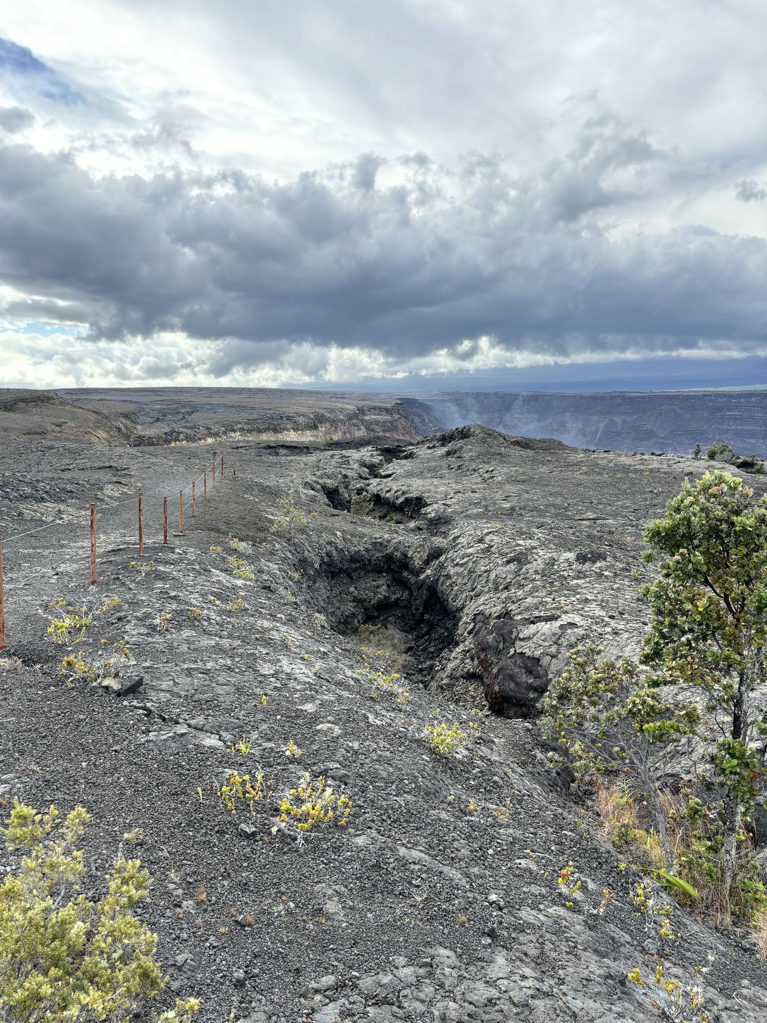 A rugged, dark volcanic landscape with deep cracks and a distant plume of smoke. - Crater Rim Drive and Devastation Trail