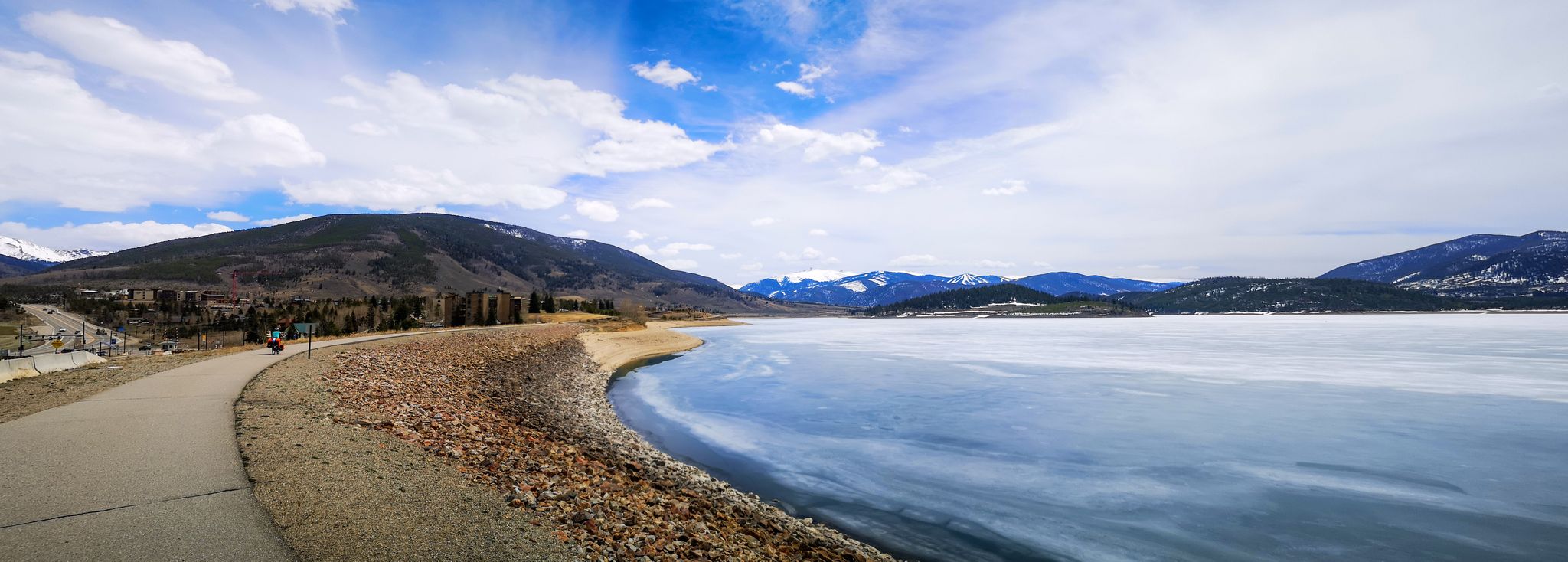 Panoramic view of a partially frozen lake, paved path, and snowy mountains. - Dillion Dam via Ryan Gulch