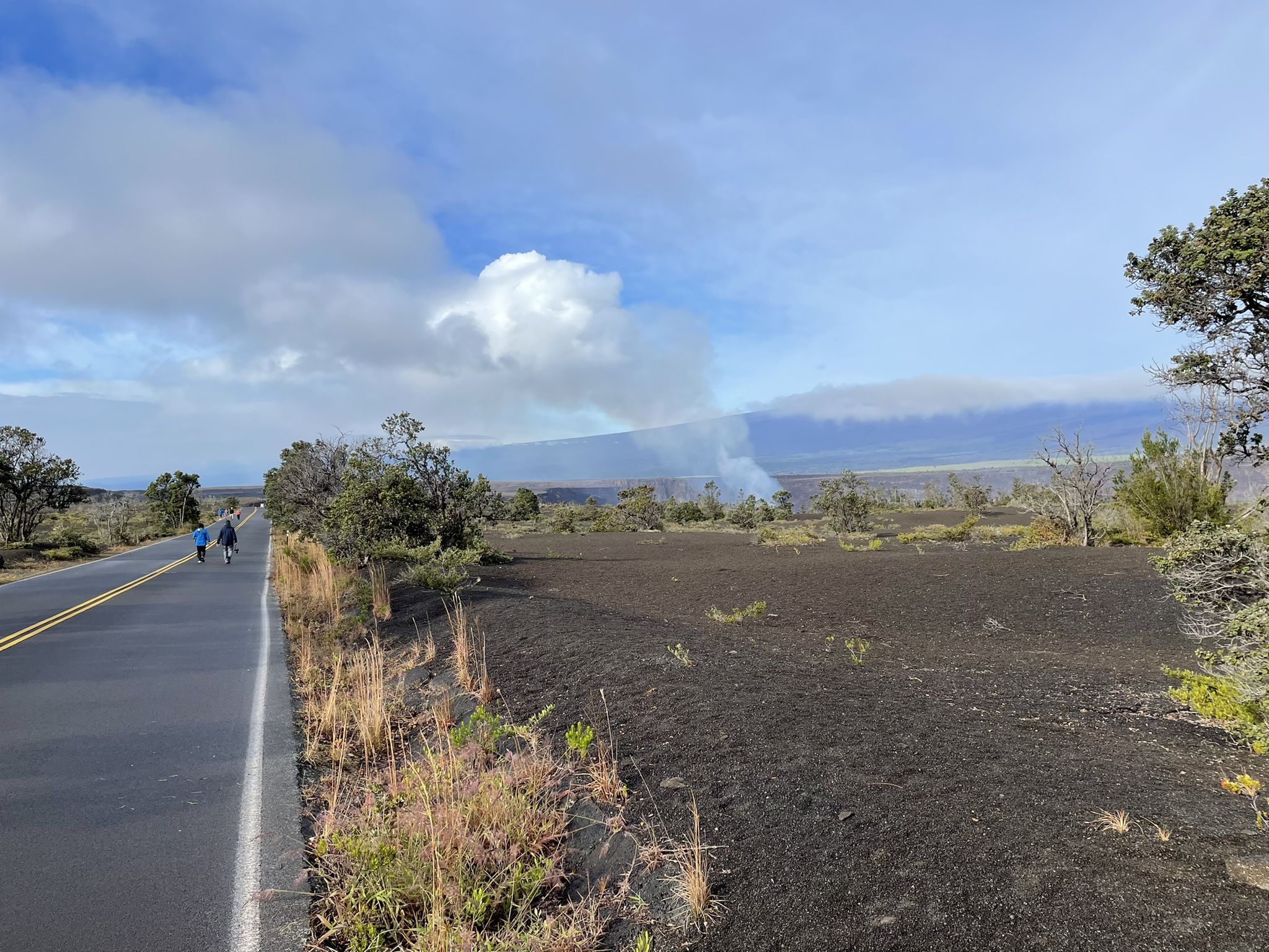 Two people walk on a road next to a dark landscape with a distant smoke plume. - Halema‘uma‘u and Keanakāko‘i Craters