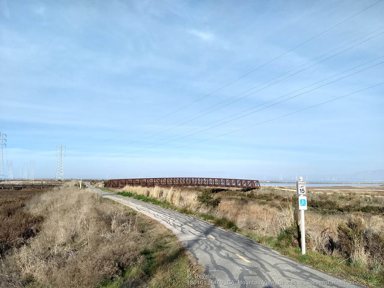 Paved road leading towards a distant metal bridge over marshland under a cloudy sky. - San Francisco Bay Trail – Moffett Boulevard Bike Bridge loop from Shoreline Amphitheatre