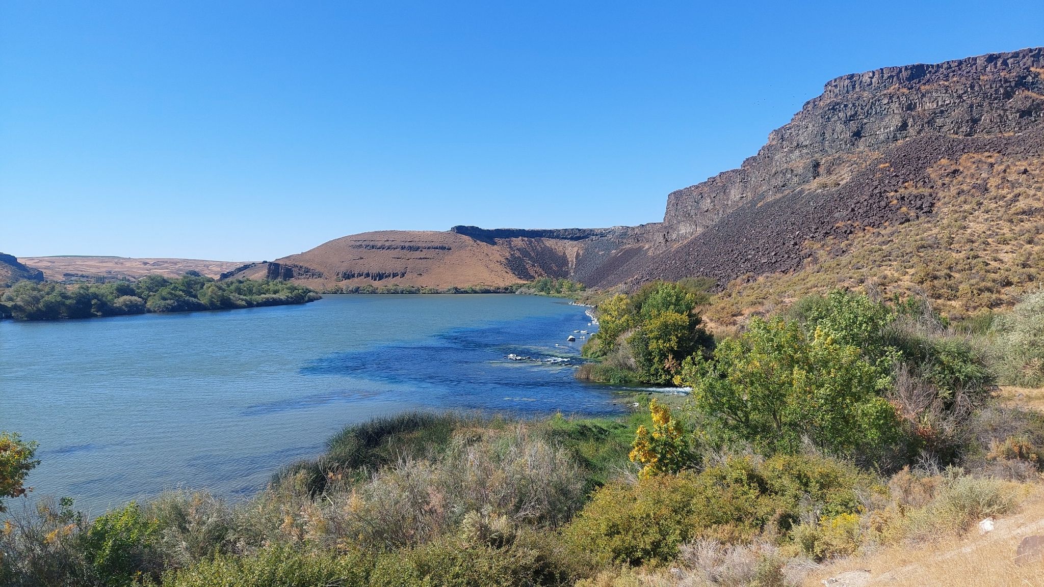 Wide river with rocky cliffs and green foliage under a clear sky. - Box Canyon Springs Trail