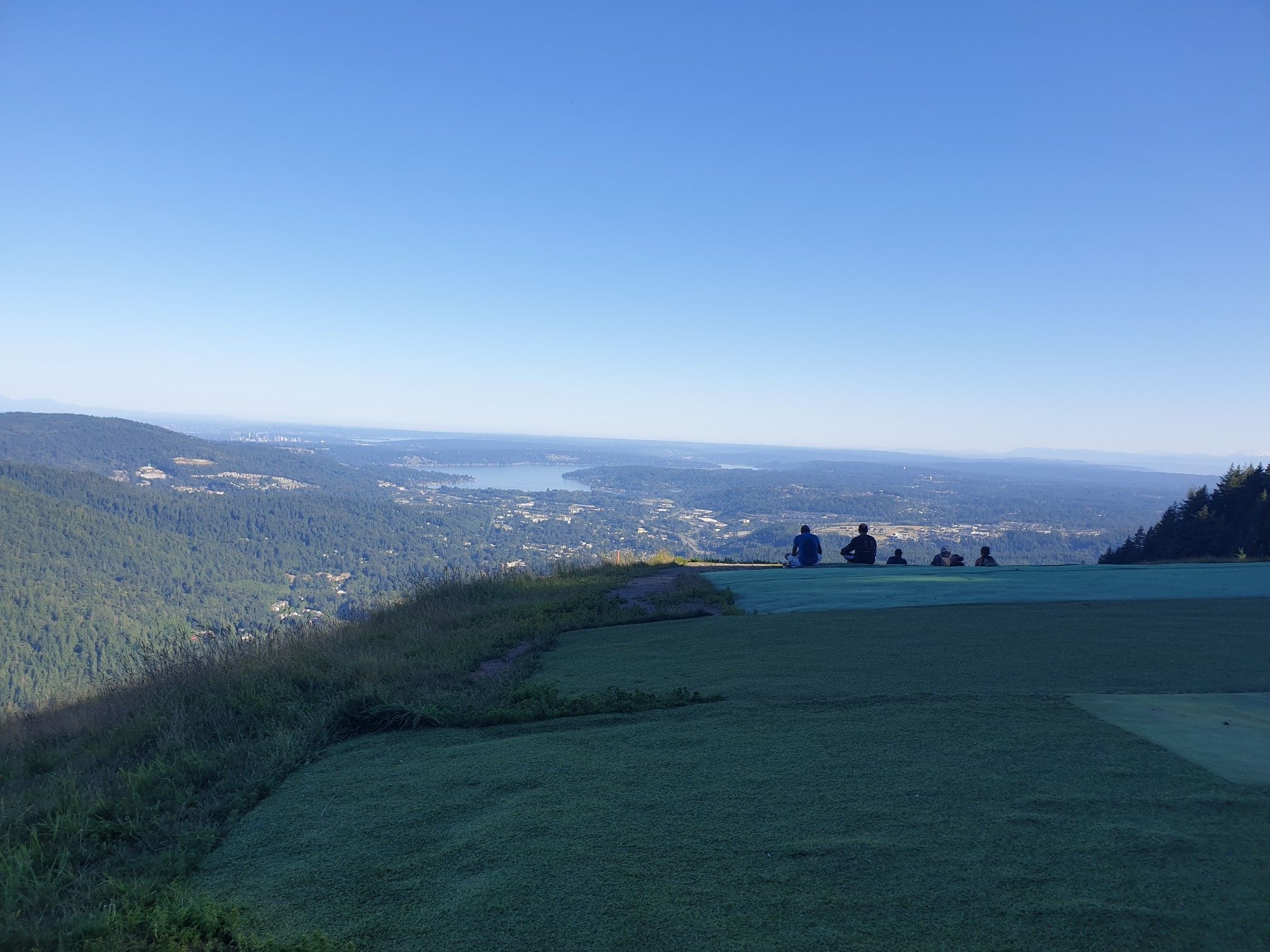 People sit on a grassy slope, overlooking a vast valley and distant water. - Poo Poo Point – Chirico Trail loop from Issaquah High School