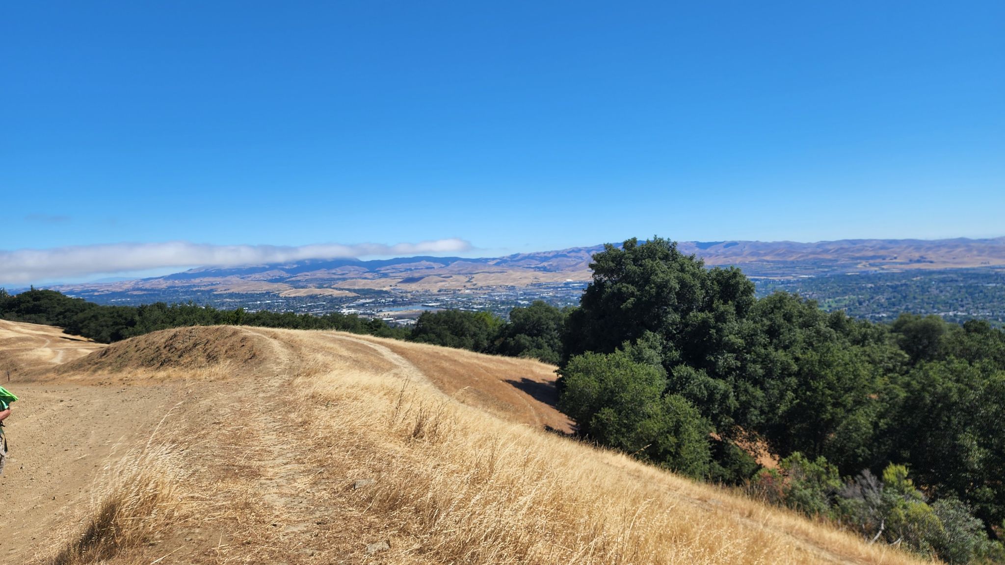 A dirt path on a golden grassy hill overlooks a distant city and mountains. - Ridgeline and Sinbad Creek Trail Loop