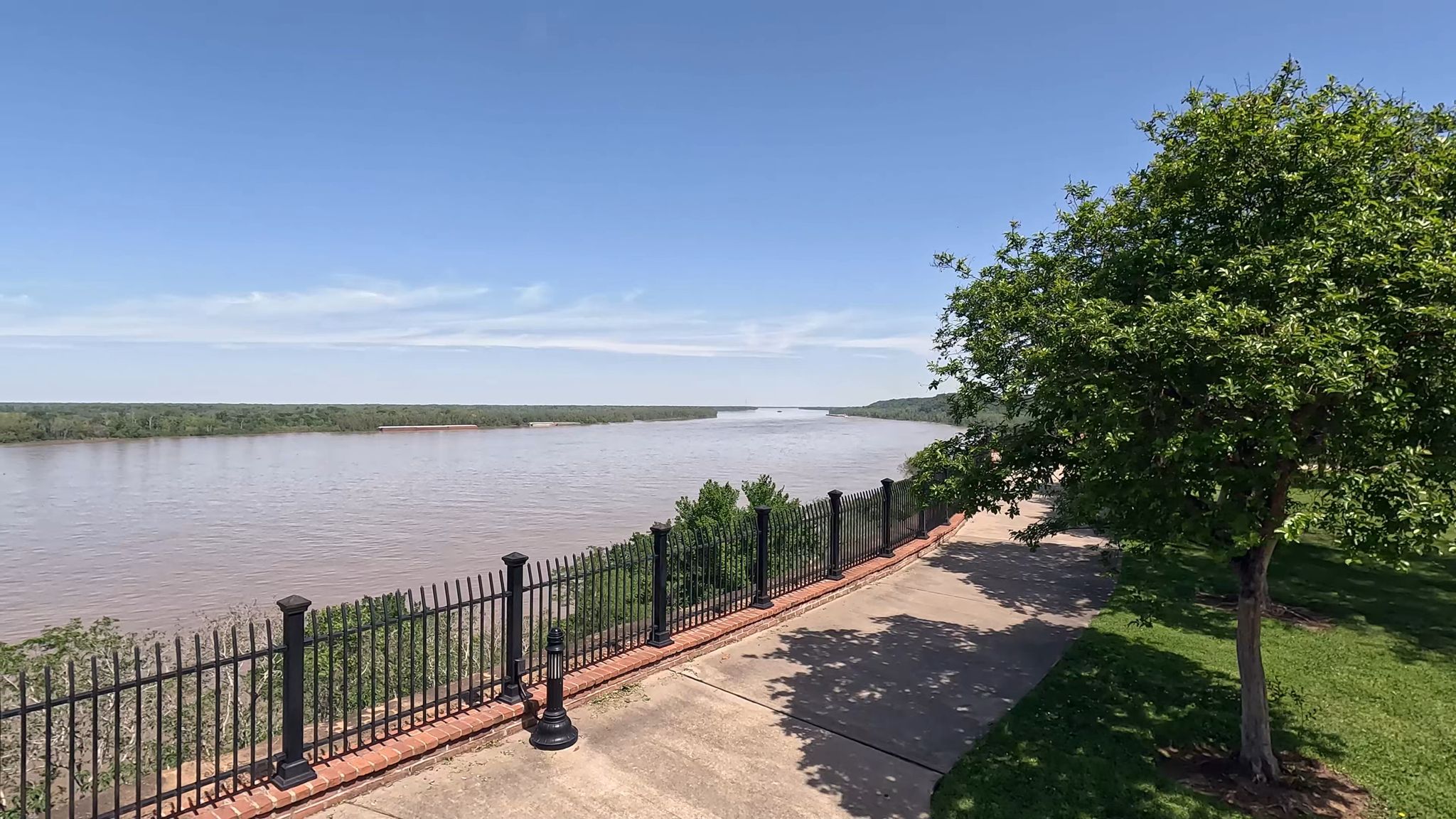 Wide river, paved path, and green trees under a bright blue sky. - Natchez Bluff Trail