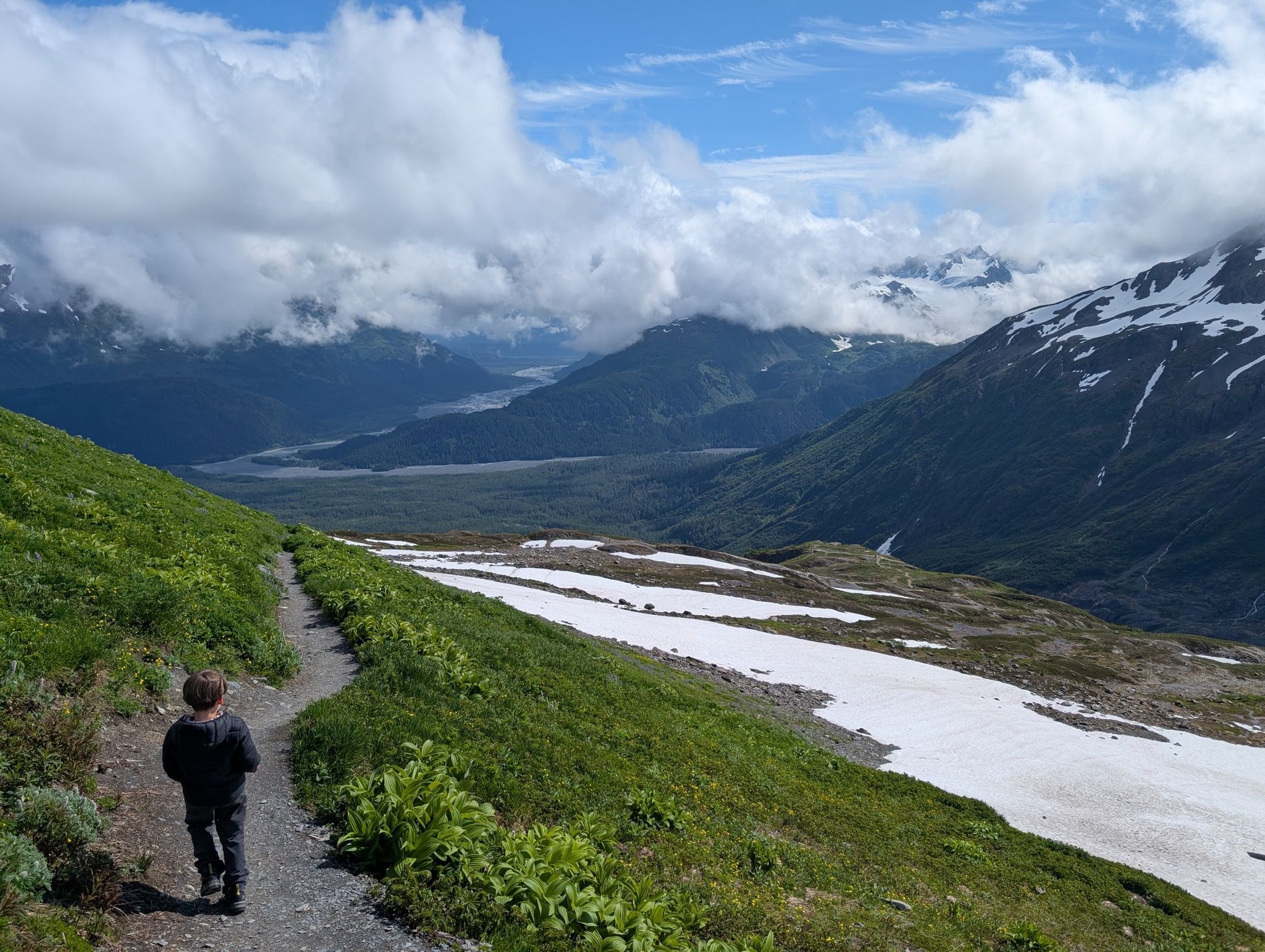 A child walks a dirt trail overlooking a valley with snow patches. - Harding Icefield Trail