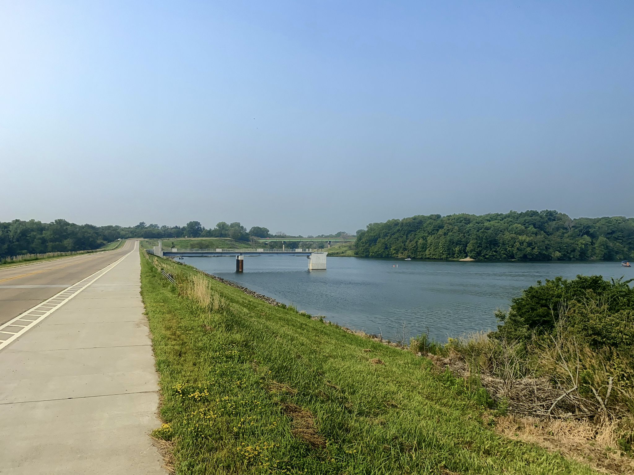 A long road runs beside a wide lake with a distant bridge. - Neal Smith Trail