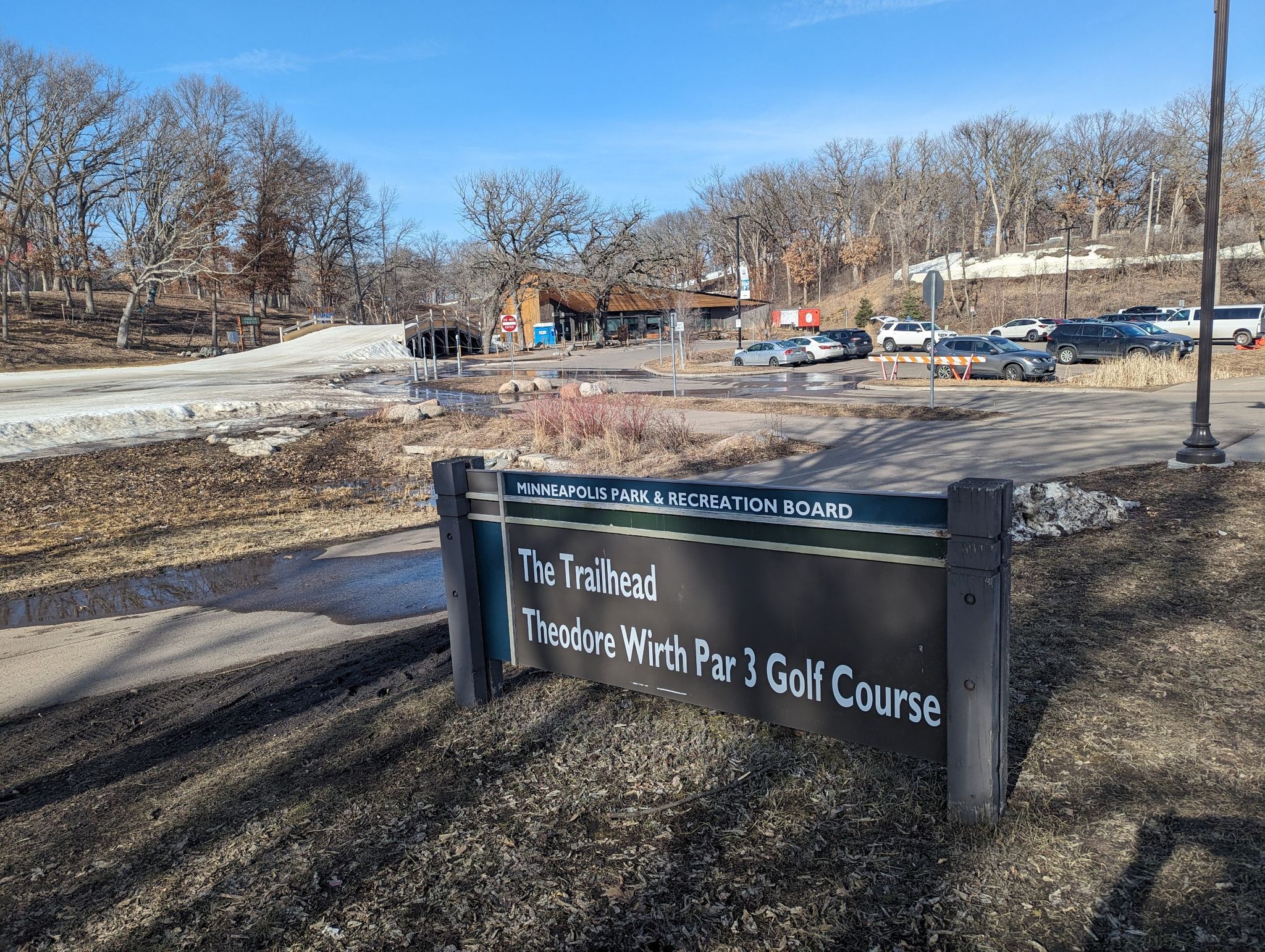 A sign in a sunny, partially snow-covered landscape with a parking lot. - Theodore Wirth Park Loop
