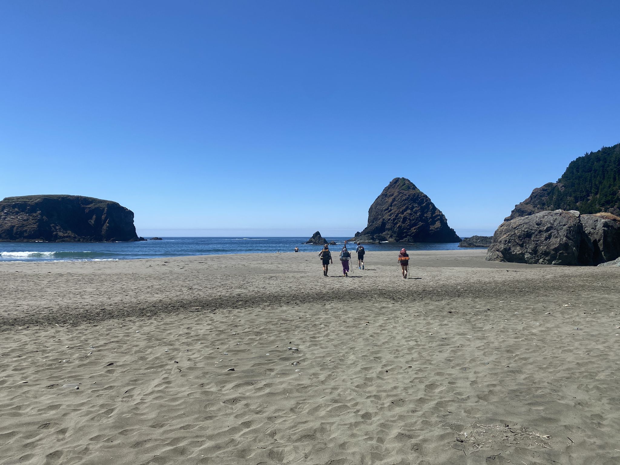 People walk on a sunny sandy beach with large rock formations and blue ocean. - Whale's Head Beach Walk