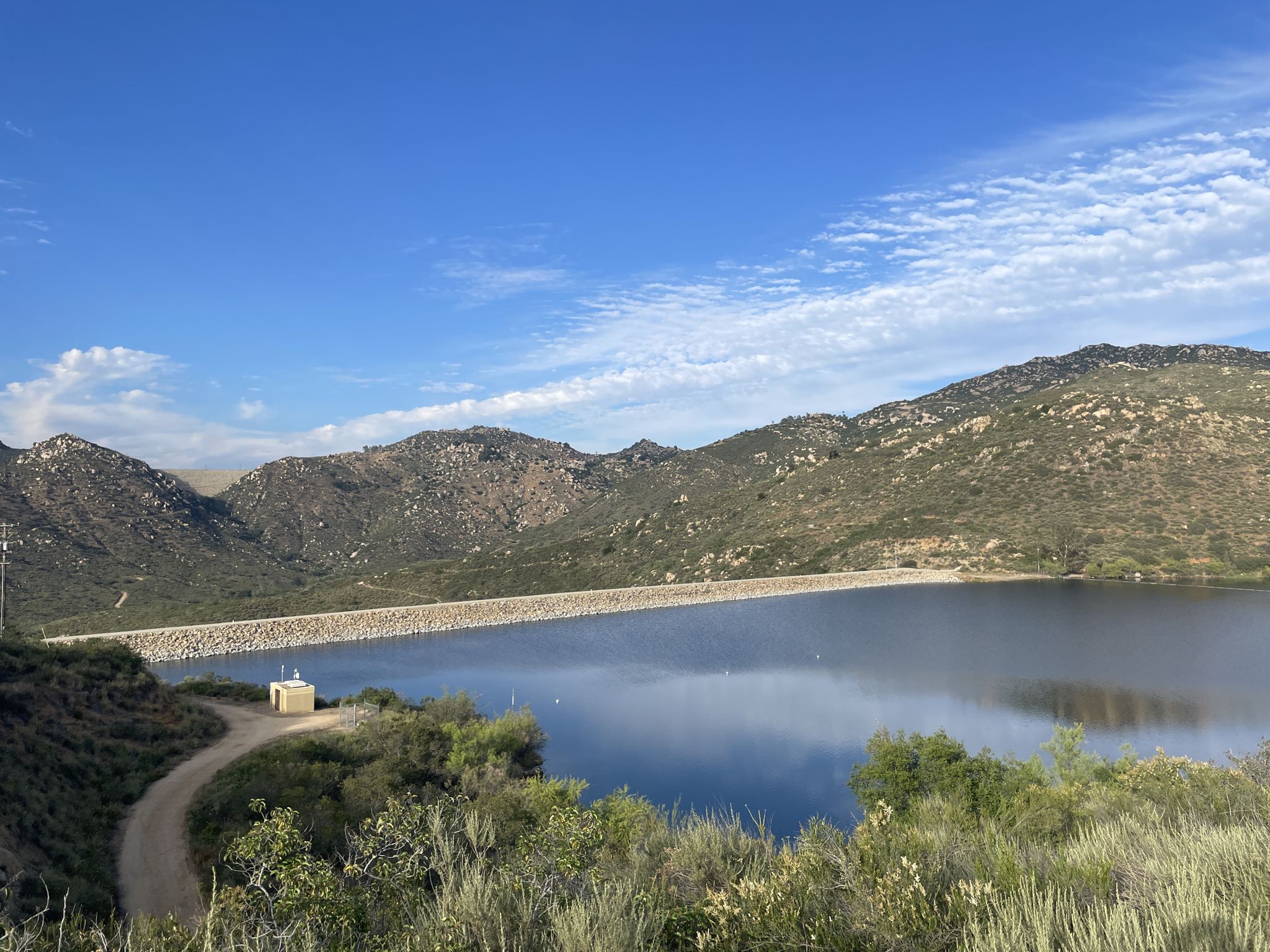 Wide lake with a dam wall, surrounded by rocky mountains under a blue sky. - Lake Poway Loop Trail
