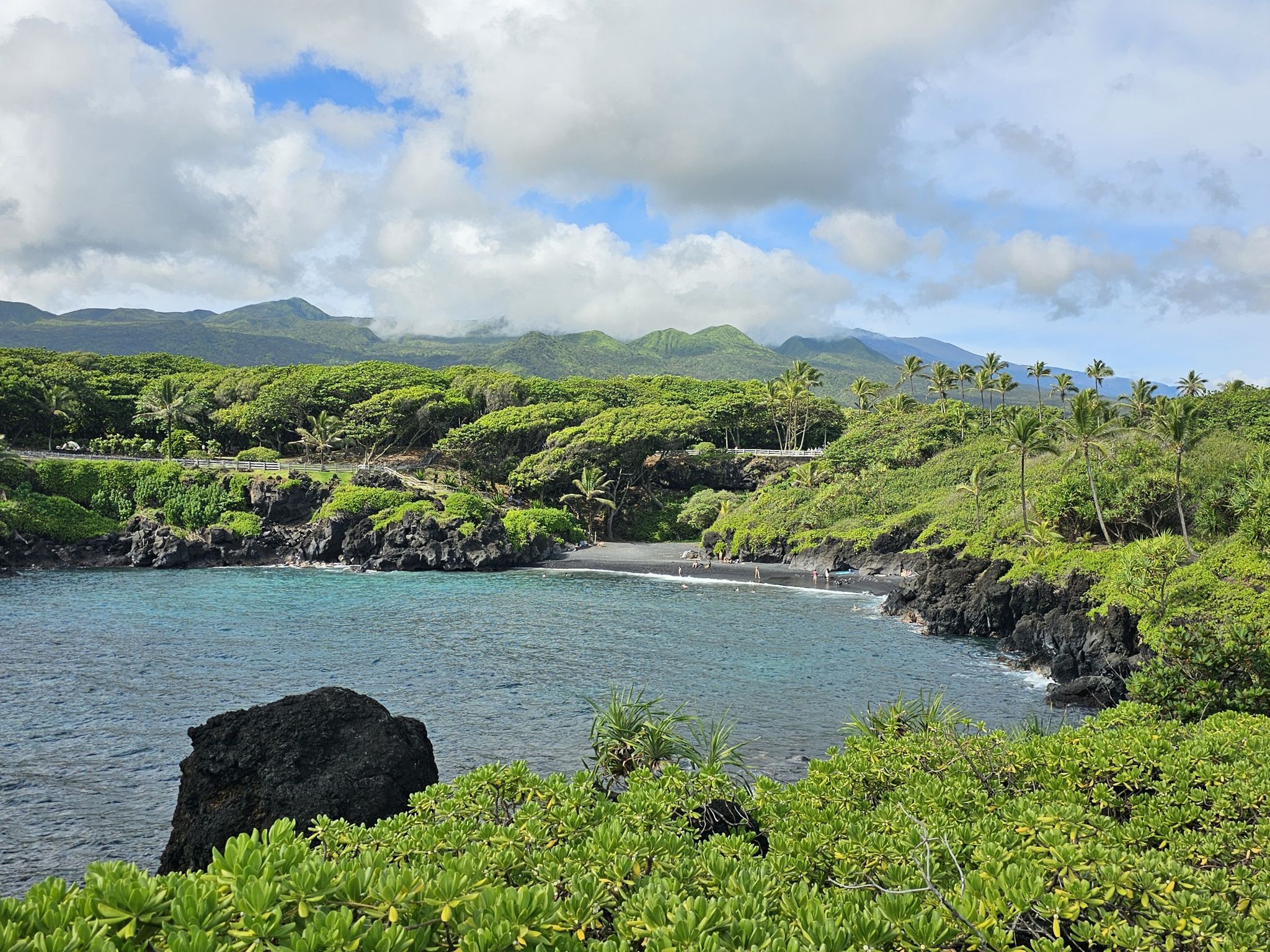 Bay with black sand beach, lush green hills, and partly cloudy sky. - Piilani Trail